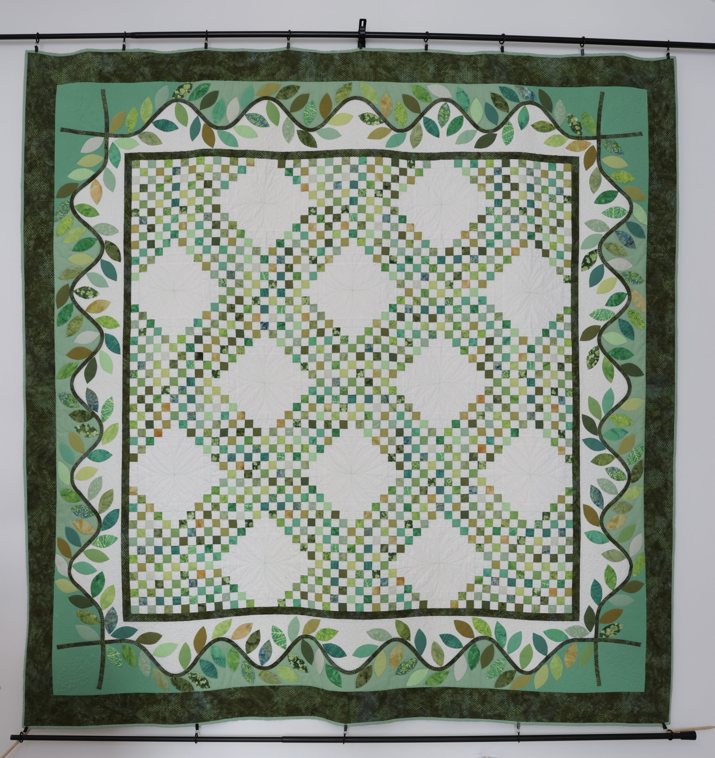A green and white quilt with a checkerboard pattern and circular designs, bordered by a leaf and vine motif.