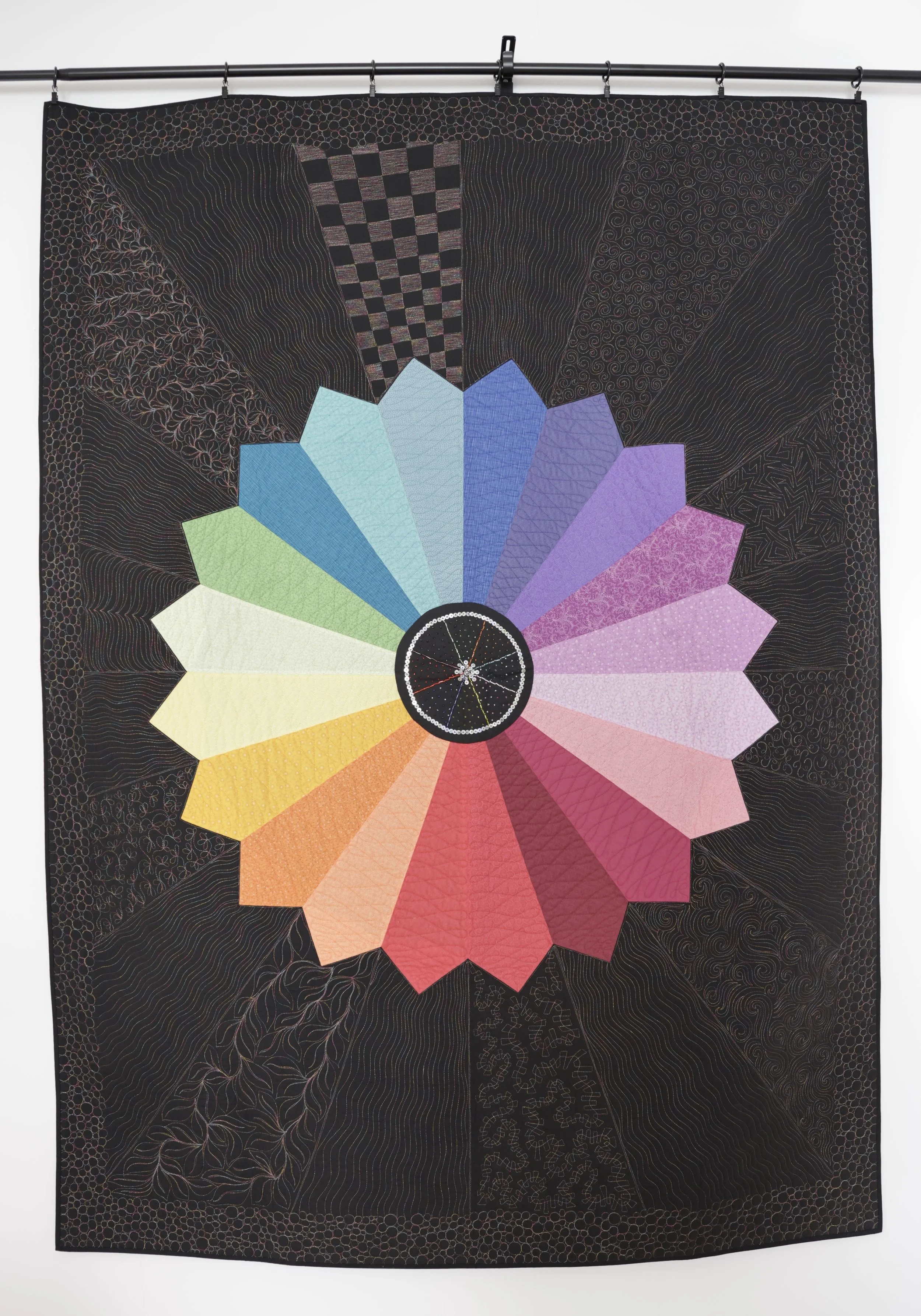 A tapestry with a black background featuring a large, circular, rainbow-colored wheel at its center. The wheel is divided into 20 segments, each in a different color, radiating outward in a gradient from red to violet. Around the wheel, intricate lin