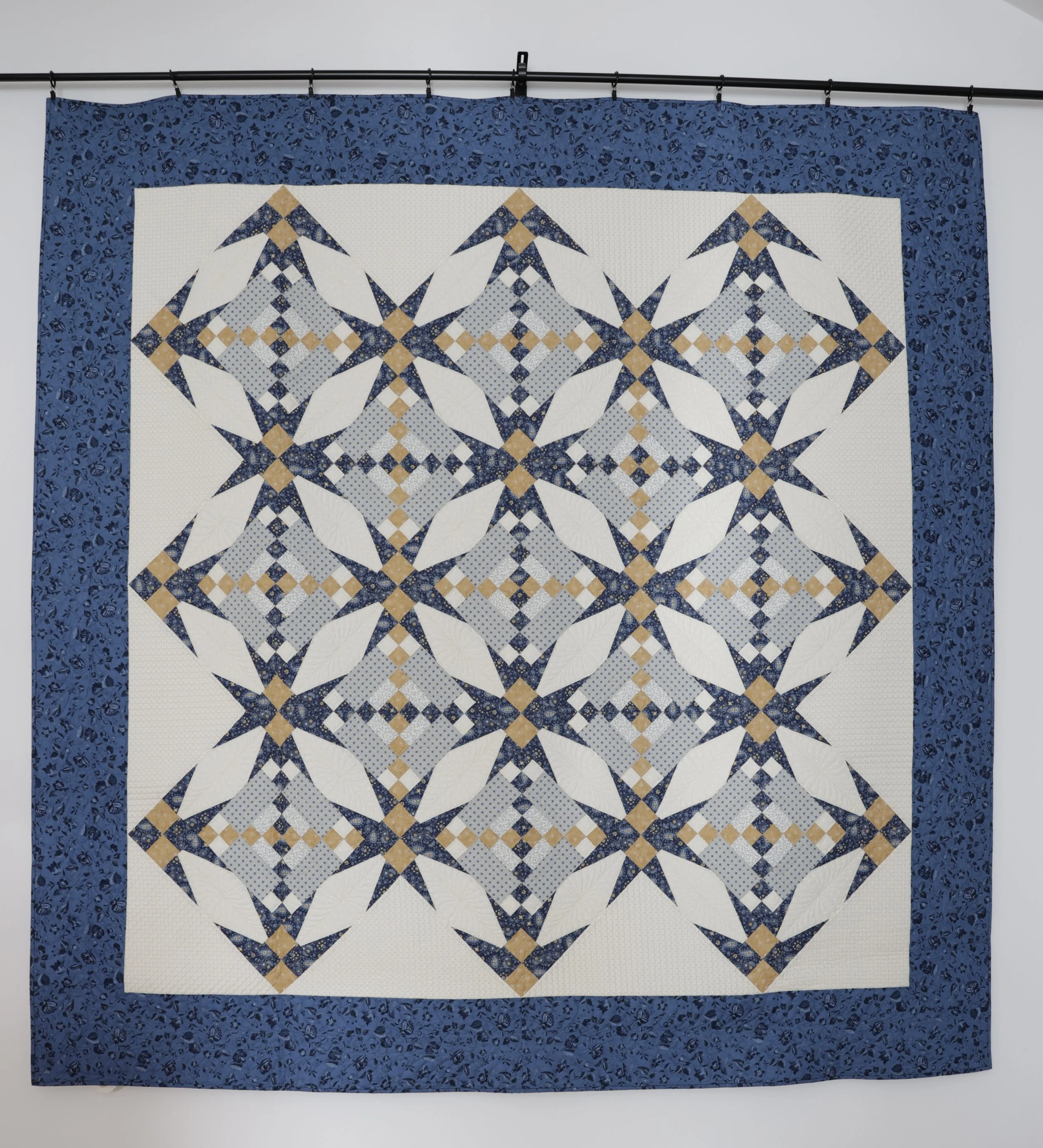 A quilt with a geometric pattern featuring navy blue, white, and tan colors, bordered by a blue patterned fabric, displayed on a wall.