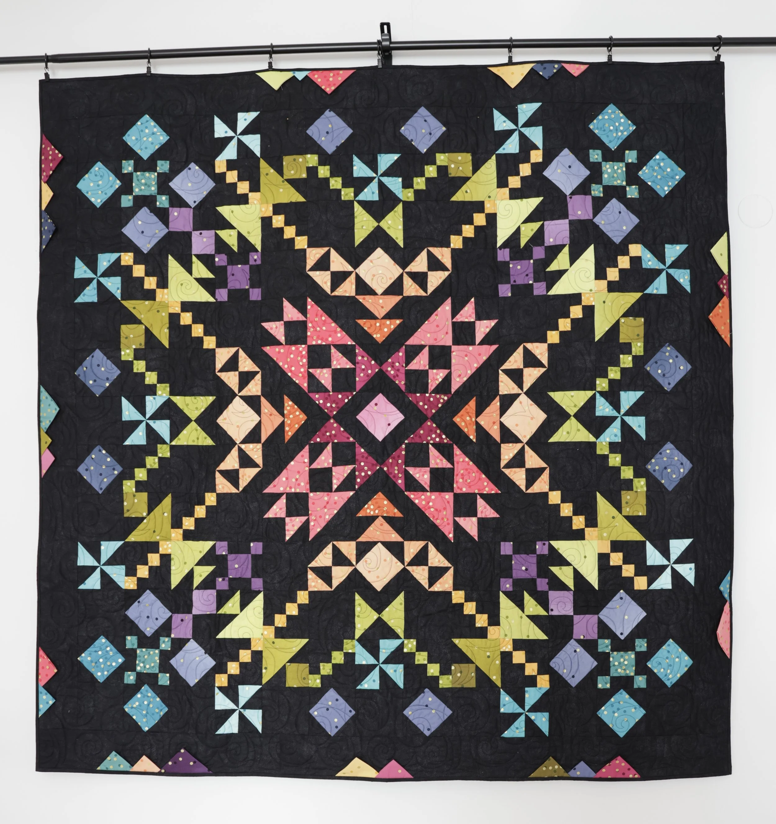 Colorful quilt with various geometric patterns on a black background, hanging on a rod.