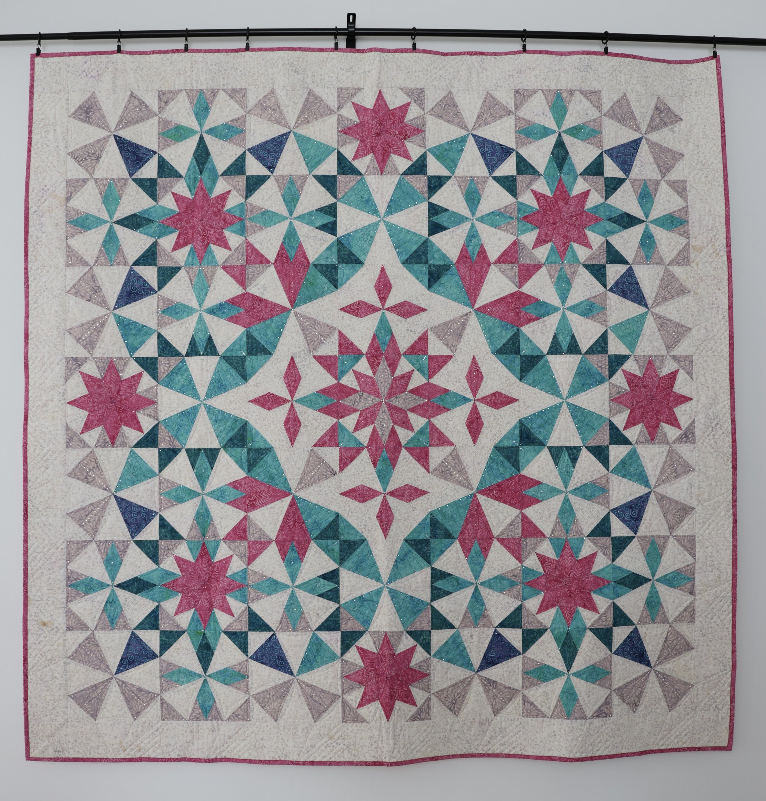 A quilt with a geometric pattern featuring pink, teal, and purple star and butterfly shapes on a white background, bordered with a pink line.
