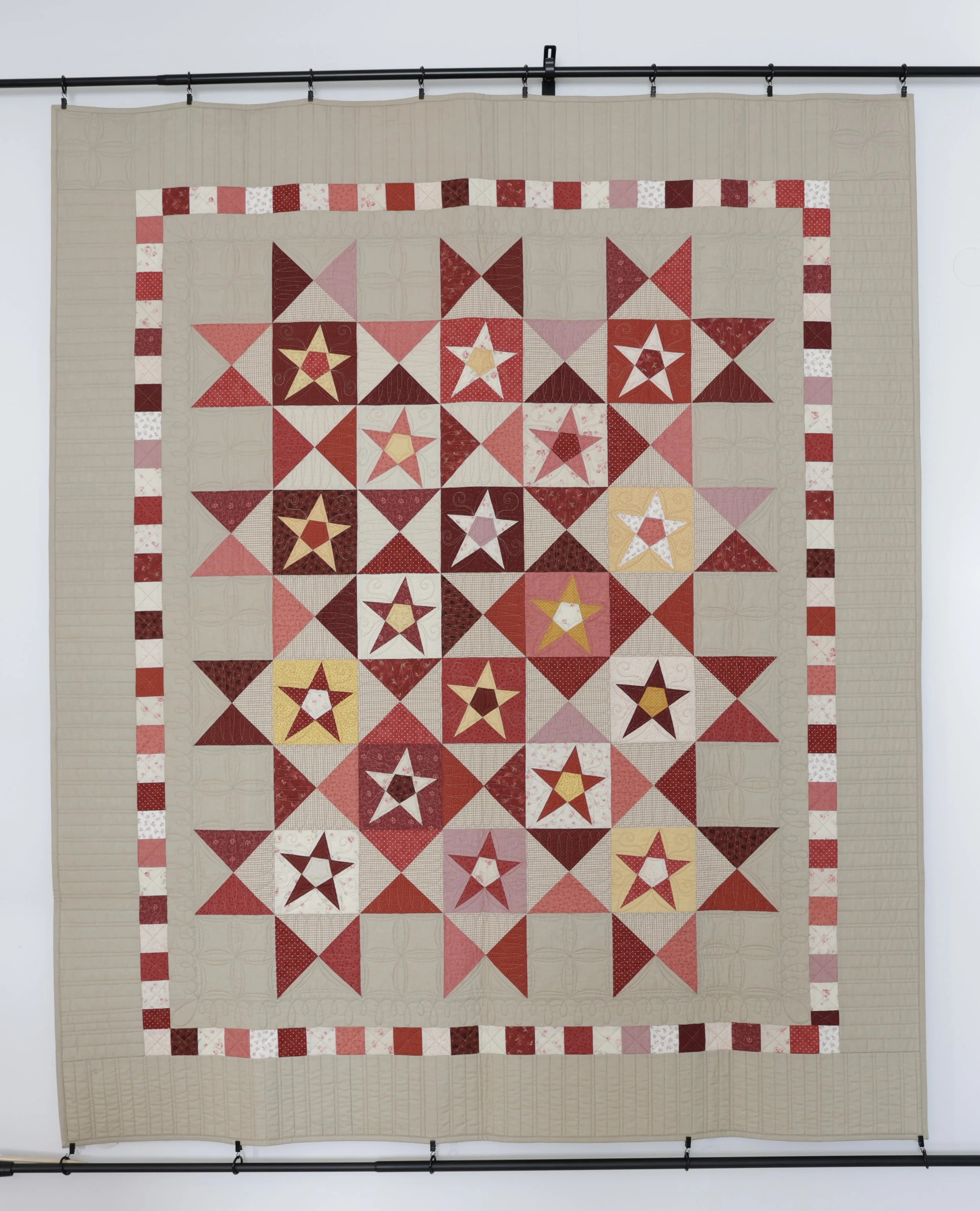 A colorful quilt with star patterns in shades of red, pink, yellow, and white, displayed on a wall with a curtain rod at the top.