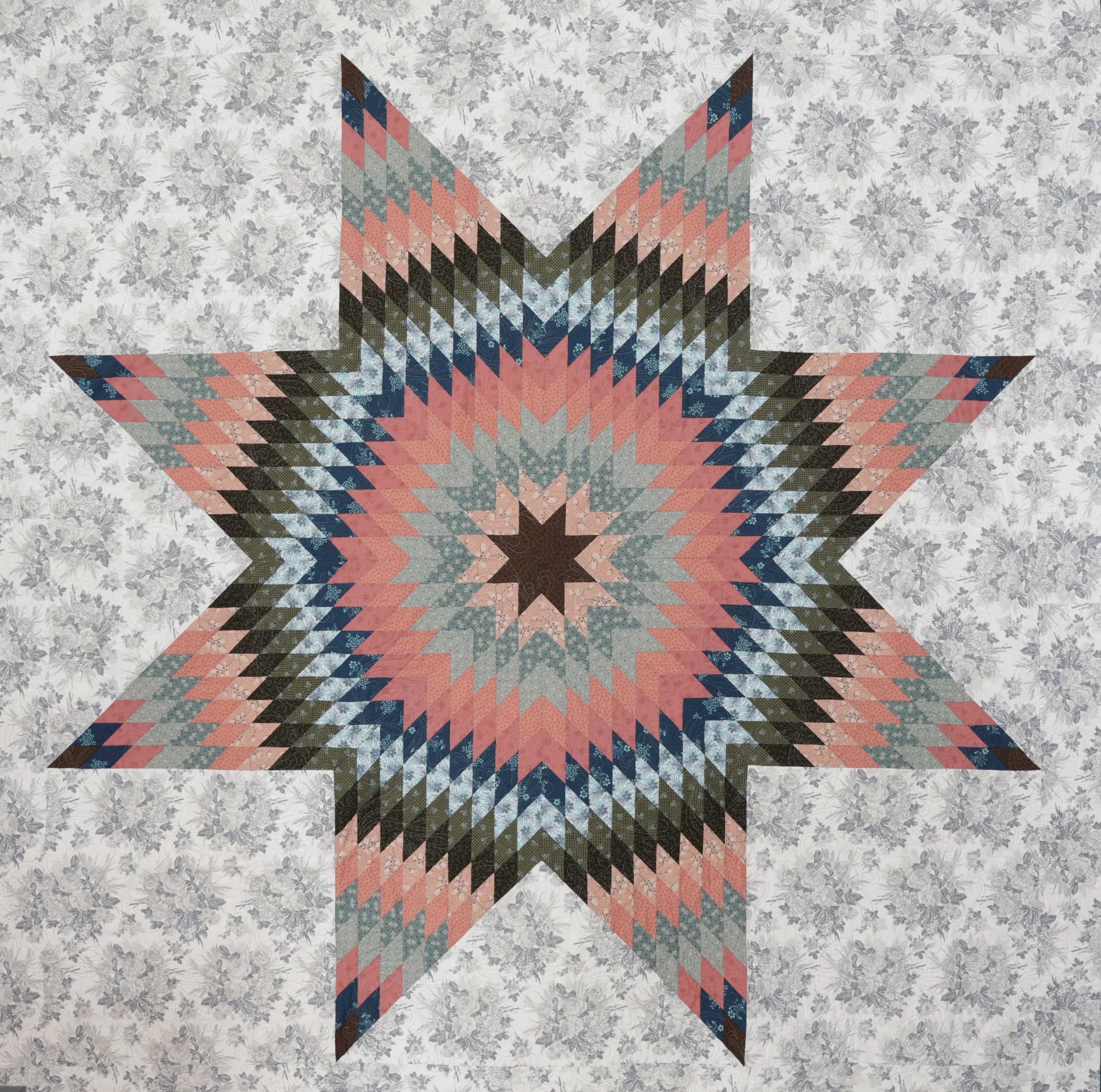 A quilt with a star pattern made of small diamond-shaped patches in pink, blue, black, and gray, on a background fabric with a gray floral print.