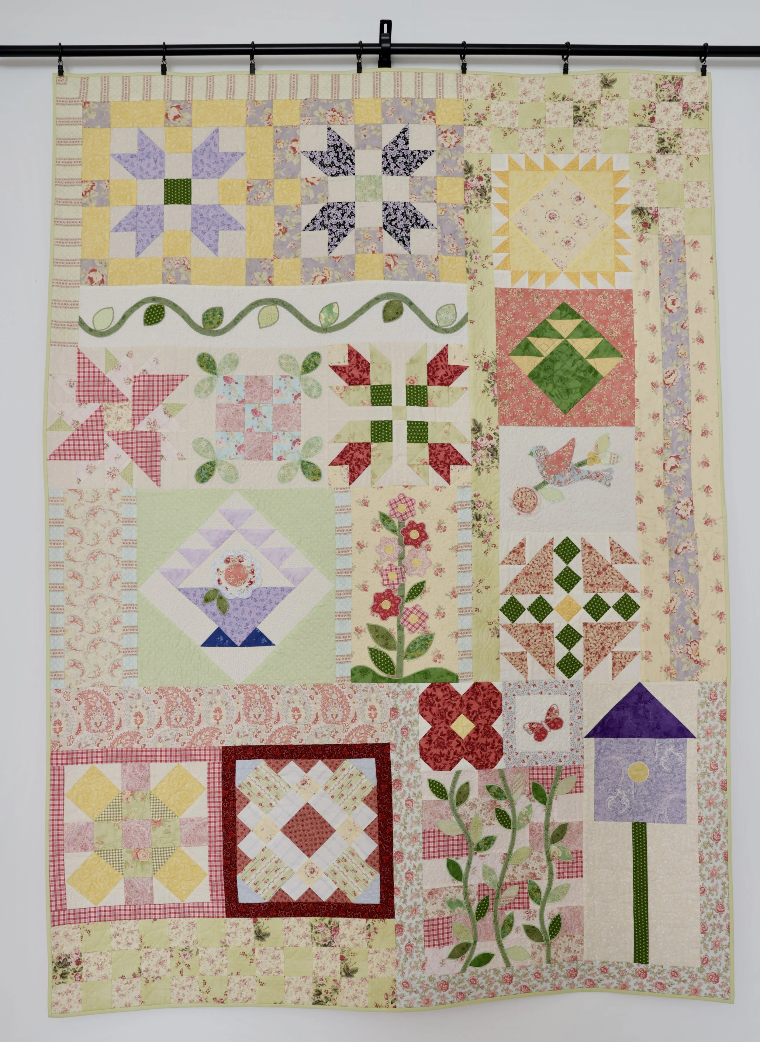 Colorful quilt hanging on a rail, featuring various patchwork and appliqué patterns including flowers, birds, houses, and geometric designs.