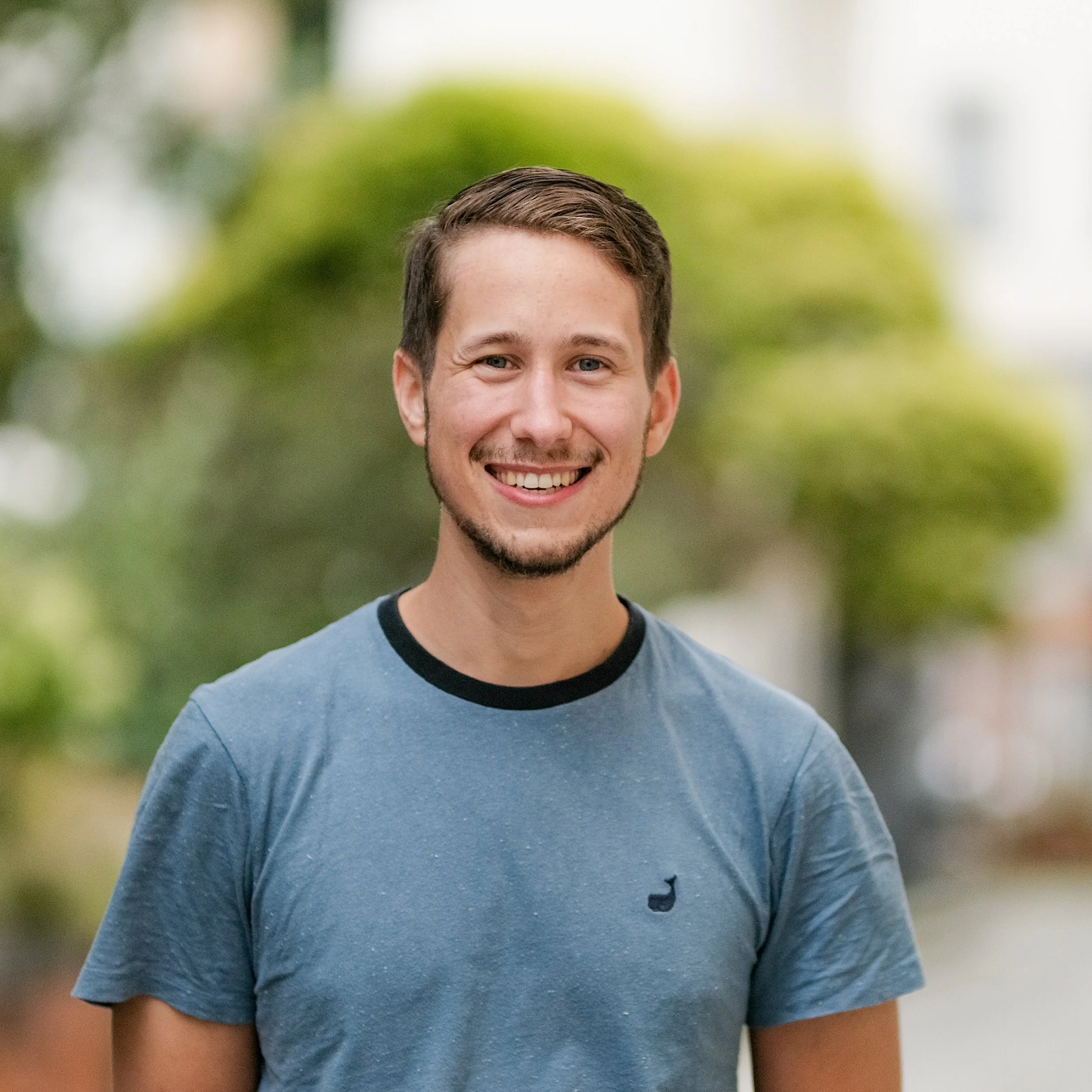 Lächelnder junger Mann mit kurzen braunen Haaren trägt ein blaugraues T-Shirt mit schwarzem Kragen und einem kleinen Logo auf der linken Brust. Er steht im Freien mit unscharfem grünen Hintergrund.