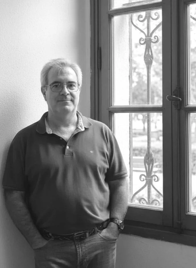 Hombre de cabello canoso, usando gafas, camiseta polo y pantalones vaqueros, apoyado en la pared junto a una ventana con rejas.