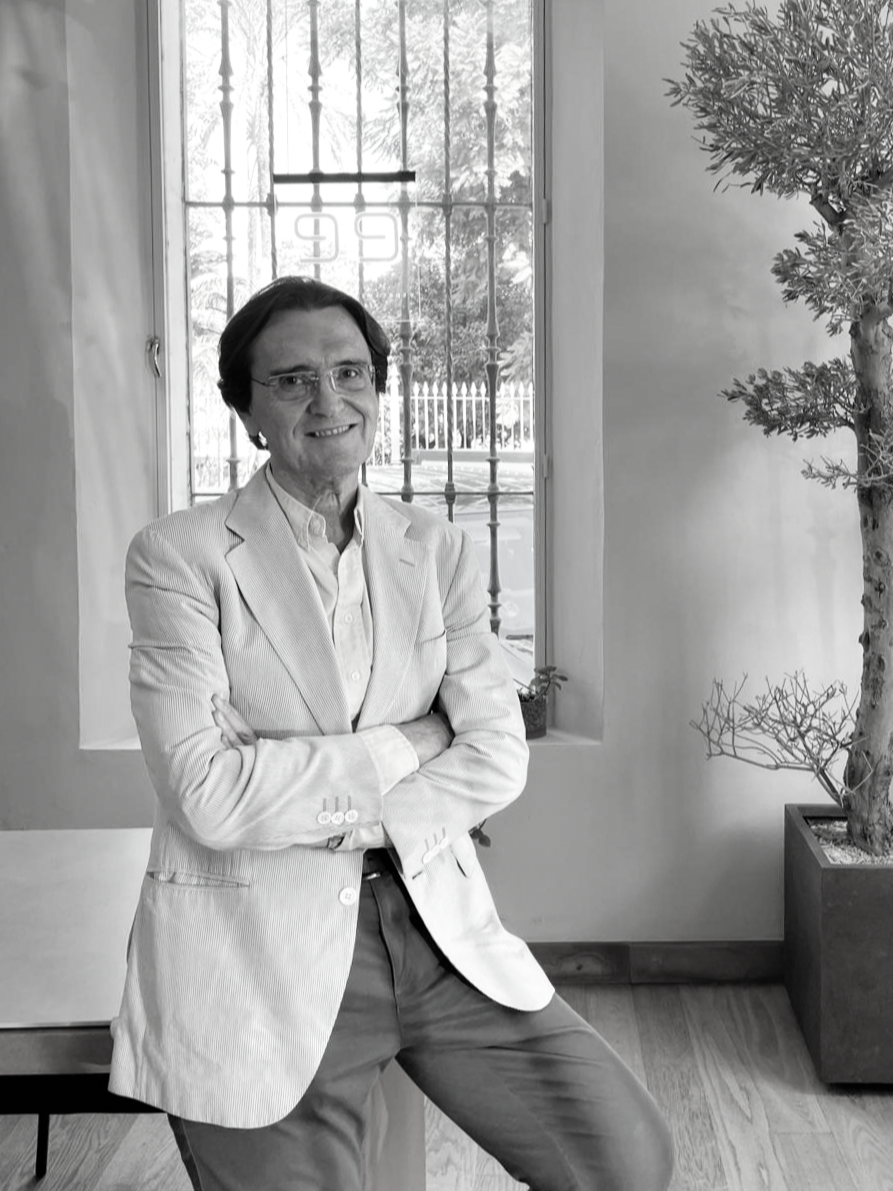 Hombre con gafas y cabello oscuro, vestido con traje claro, sonriendo, de pie en interiores cerca de una ventana con barrotes, con plantas en macetas a su alrededor.