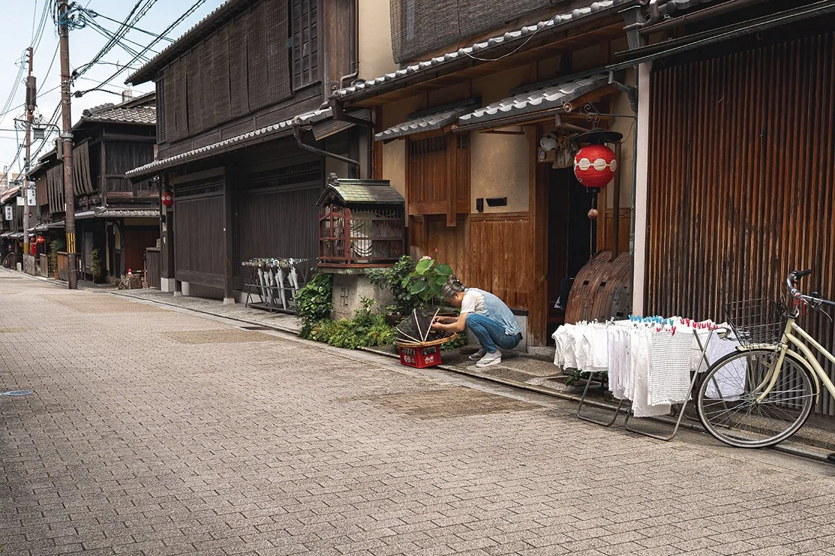 Une rue traditionnelle japonaise avec une maison en bois et des objets suspendus. Une femme plie du linge blanc à l'extérieur, à côté d'un vélo. La rue est calme et pavée, avec des câbles électriques au-dessus.