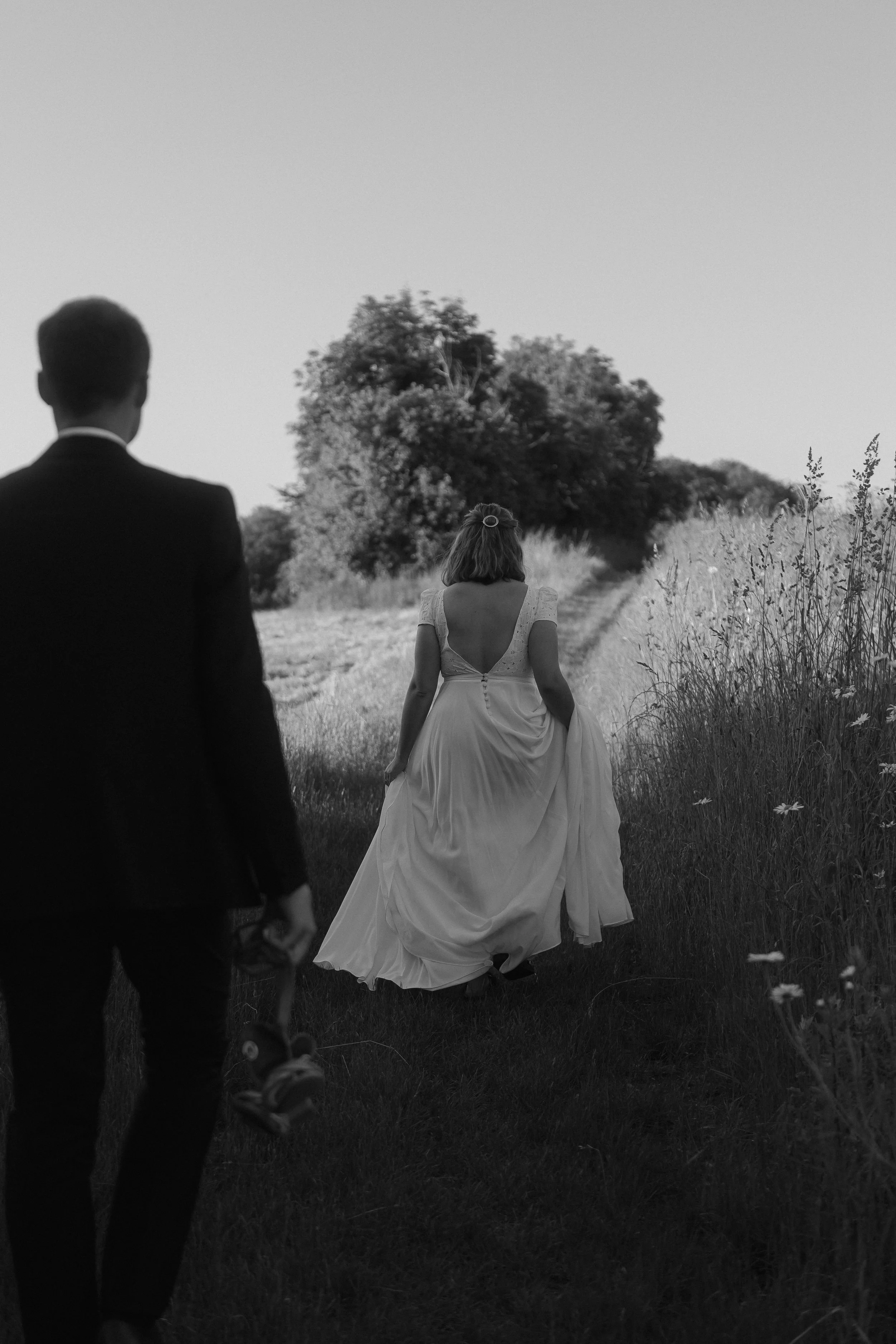 Une femme en robe de mariage marche dans un champ du Val d'Oise, le marié en costume la suit, dans un paysage naturel avec des arbres et des fleurs sauvages.