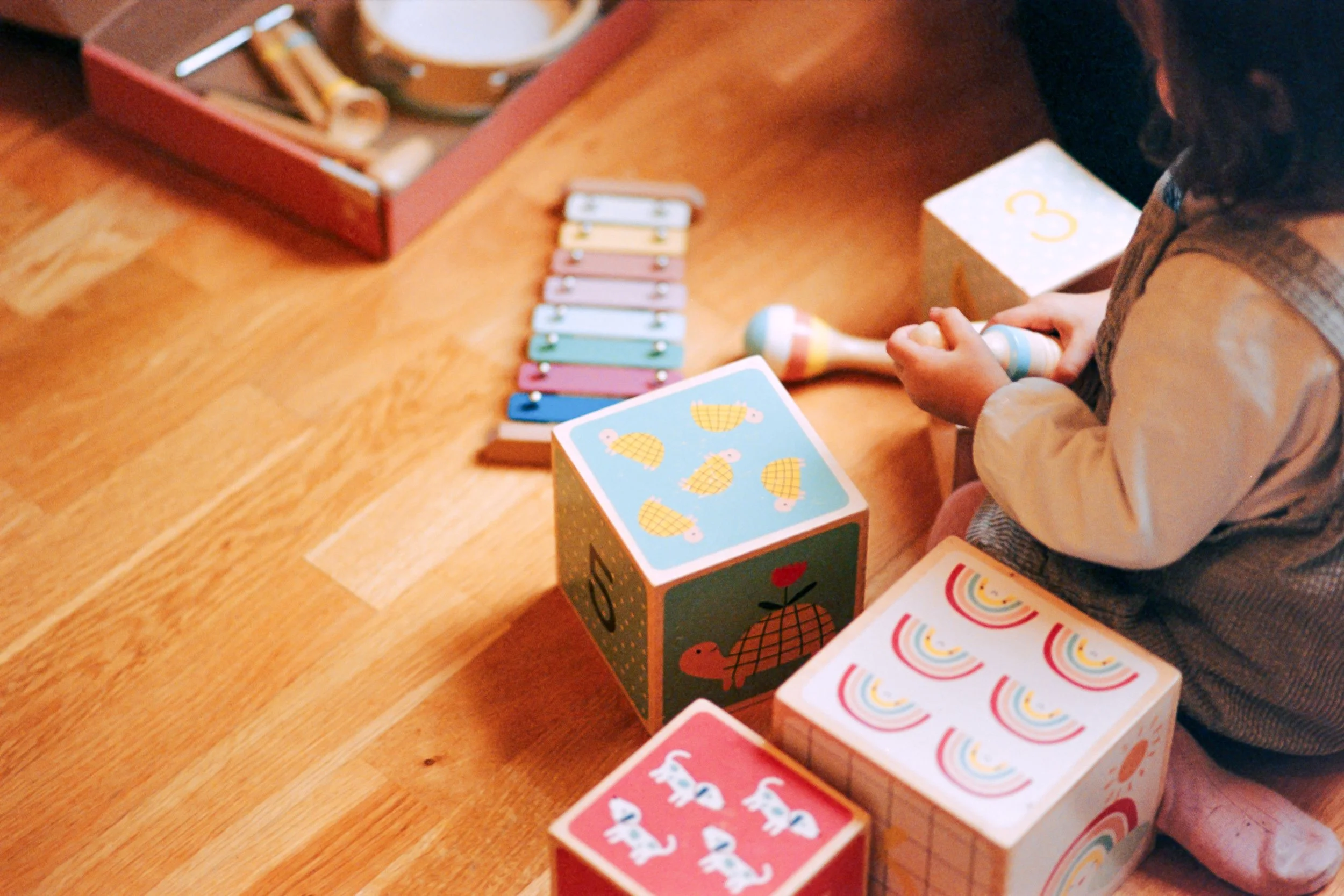 Une petite fille jouant avec des cubes colorés à motifs de canards, arcs-en-ciel, lapins et autres dessins, sur un plan en bois, avec un jeu de cartes et une boîte de jouets à proximité.