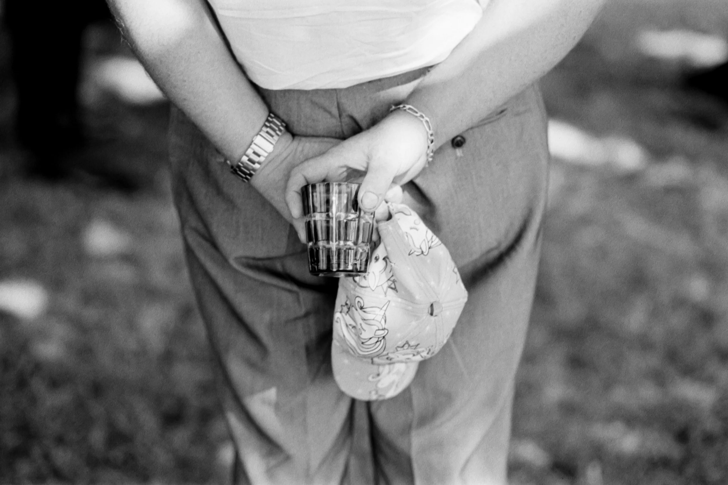 Une personne à un cocktail de mariage portant une chemise blanche, un bracelet en métal, et un bracelet chaîne, tenant un verre et un chapeau à la main, en noir et blanc. Un aspect poétique et nostalgique.