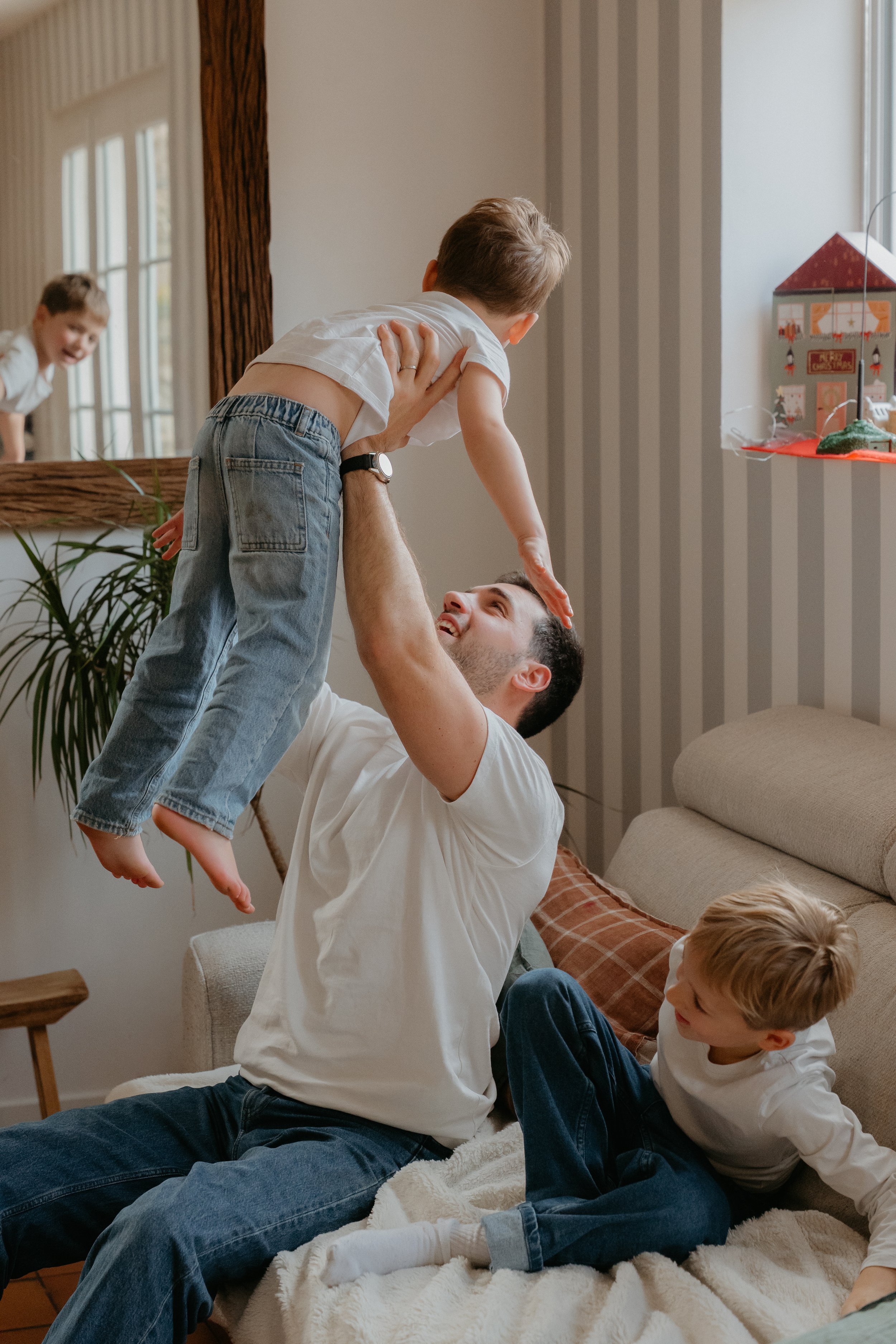 Un père des Yvelines joue avec ses enfants dans un salon lors d'une séance photo famille, l'un d'eux étant levé en l'air par le père, l'autre regardant et souriant.