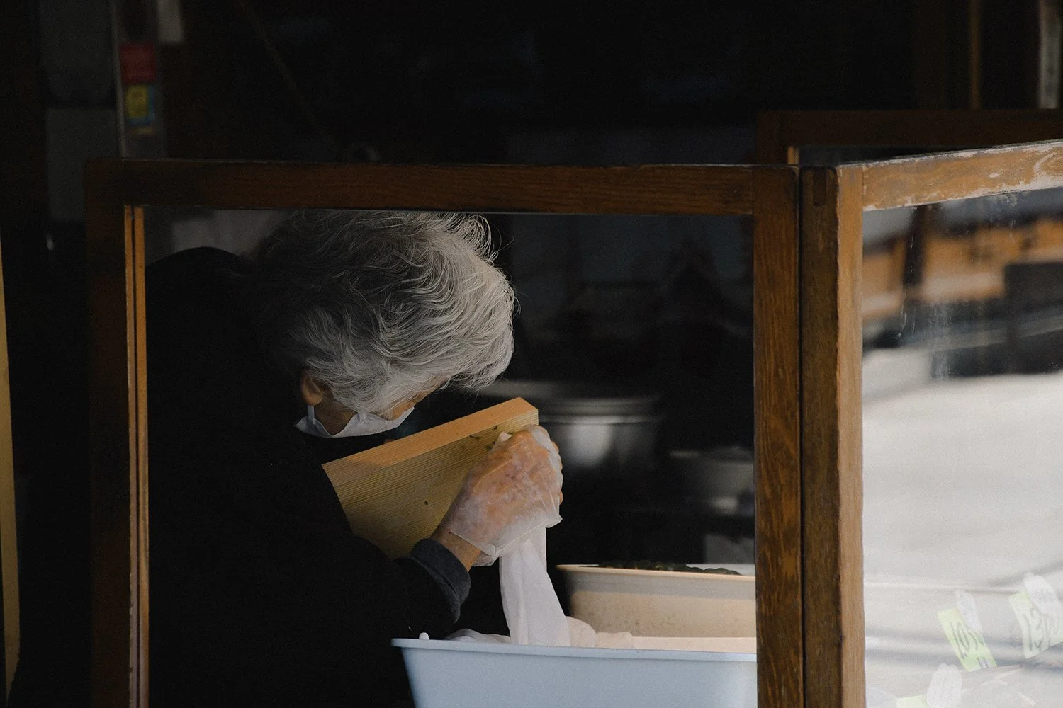 Une vieille femme aux cheveux gris travaillant derrière une vitrine en bois, manipulant un objet dans un établissement de nourriture ou un marché.