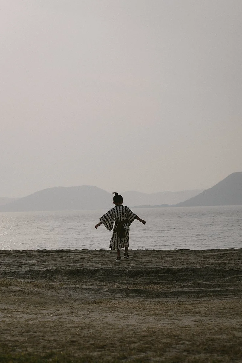 Une personne portant un kimono ou un vêtement traditionnel japonais, marchant ou courant vers la mer, avec des montagnes en arrière-plan, au coucher ou au lever du soleil.