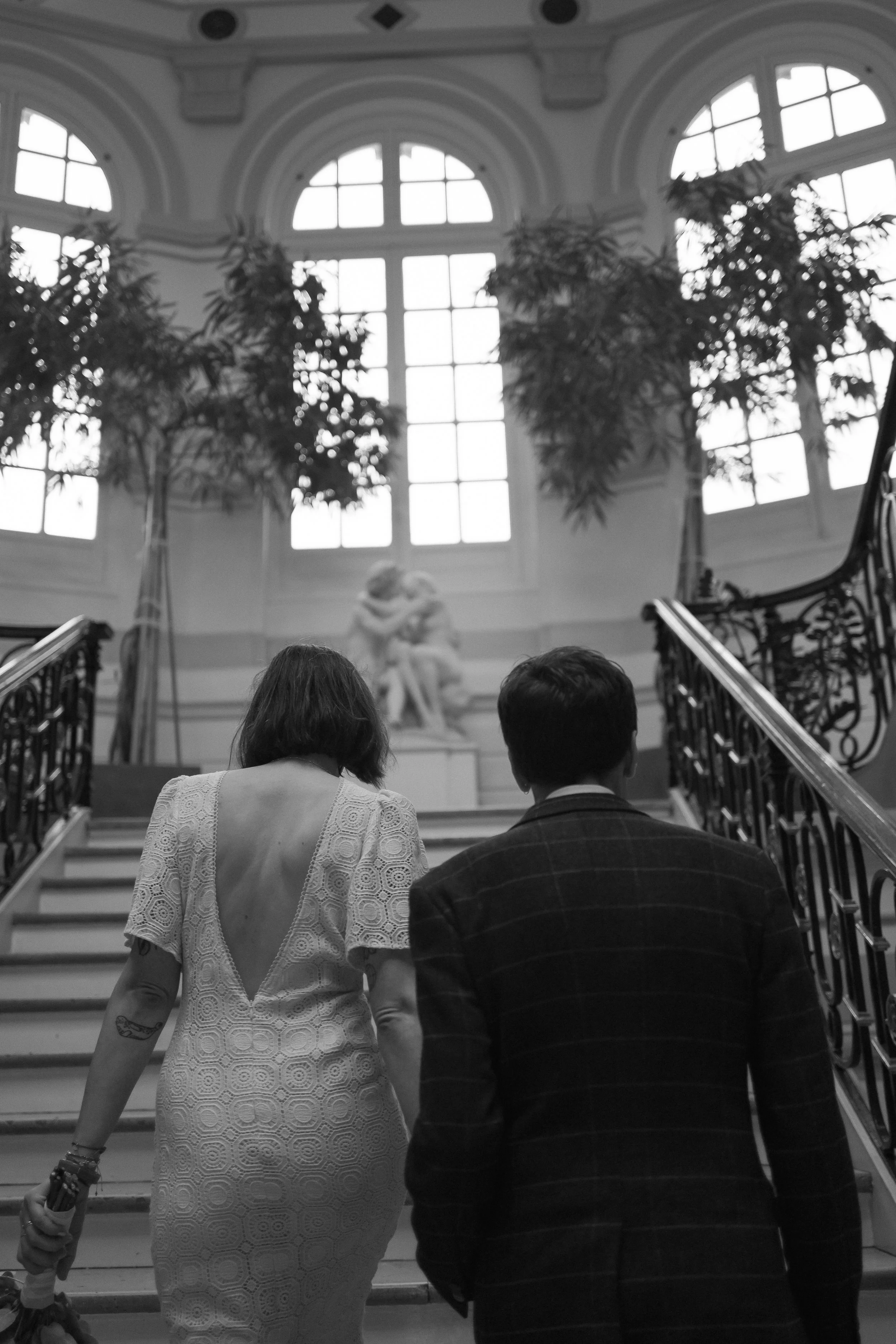 Un couple de mariés monte des escaliers dans une salle élégante avec de grandes fenêtres et une sculpture au fond.