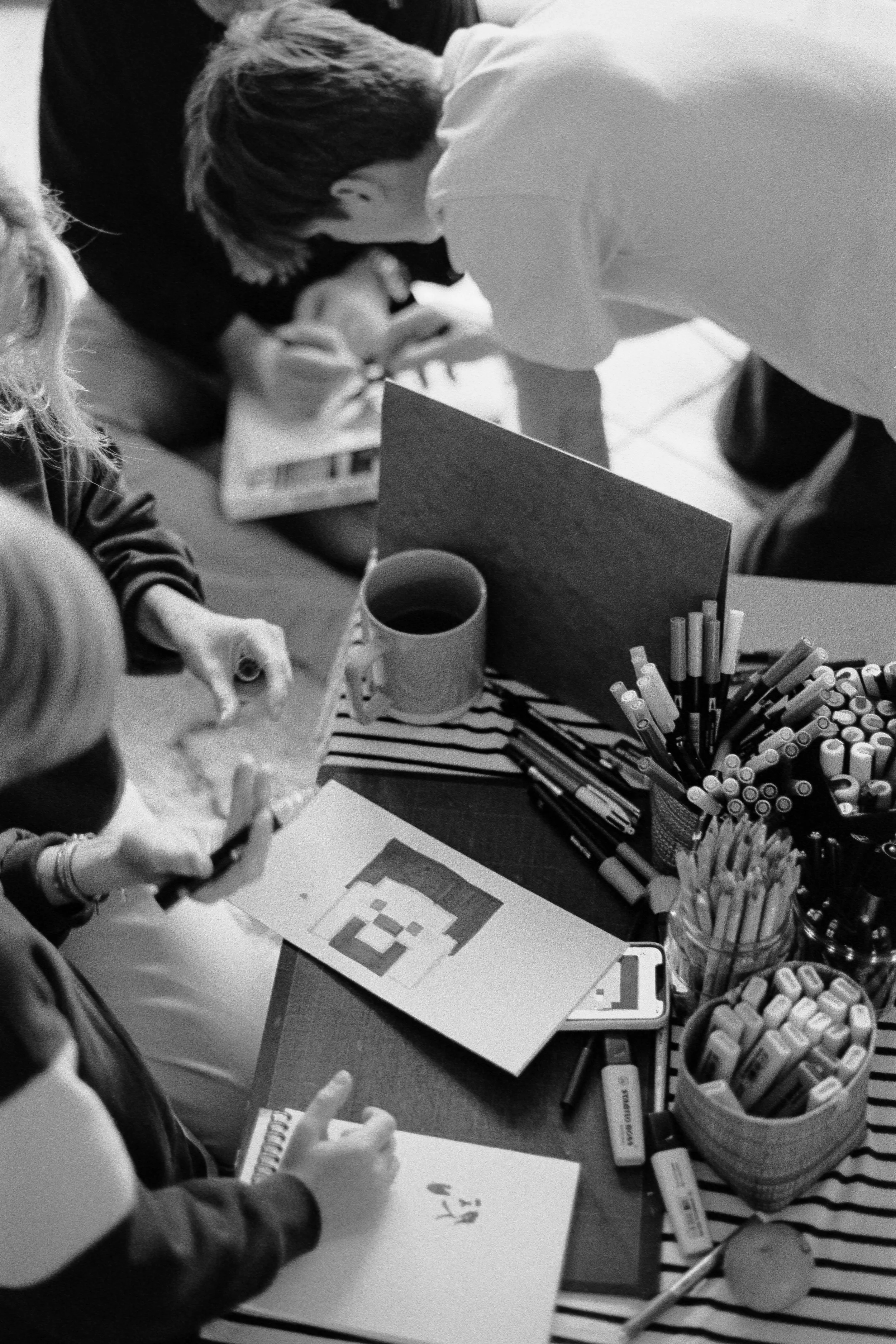 Famille autour d'une table avec des fournitures d'art, un ordinateur portable, des tasses de café, et des dessins. Le tout en noir et blanc.