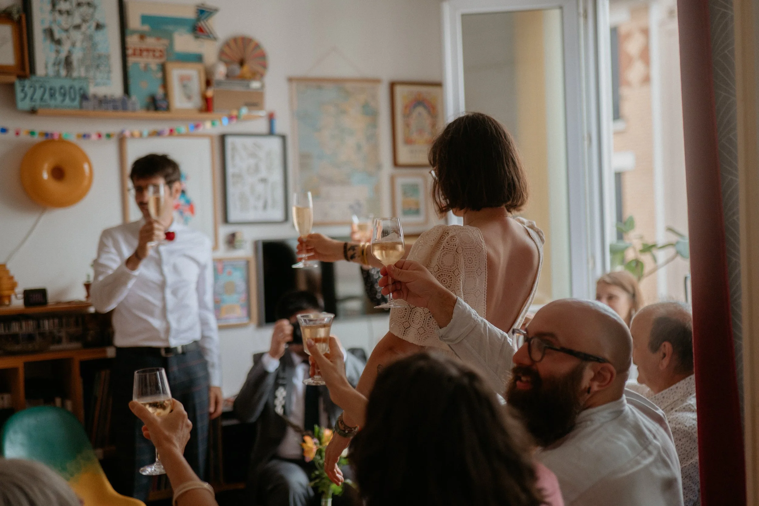 Une famille trinque à un mariage, tenant des verres de champagne, dans une pièce décorée avec des tableaux, des guirlandes et des objets colorés.