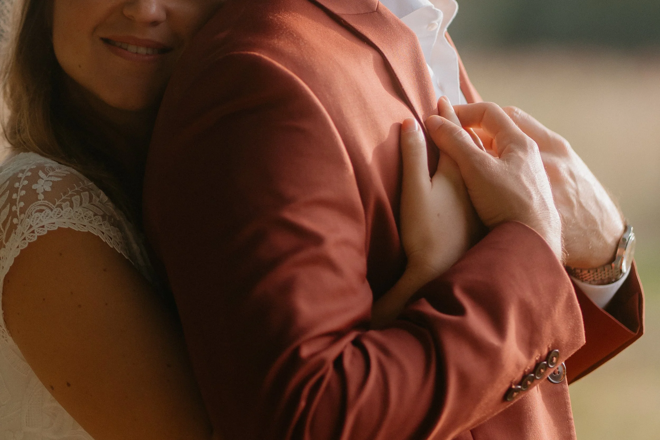 Un jeune marié porte un costume de mariage en terracota et tient les bras de la mariée en robe de mariage. La scène semble être un câlin ou une étreinte chaleureuse.