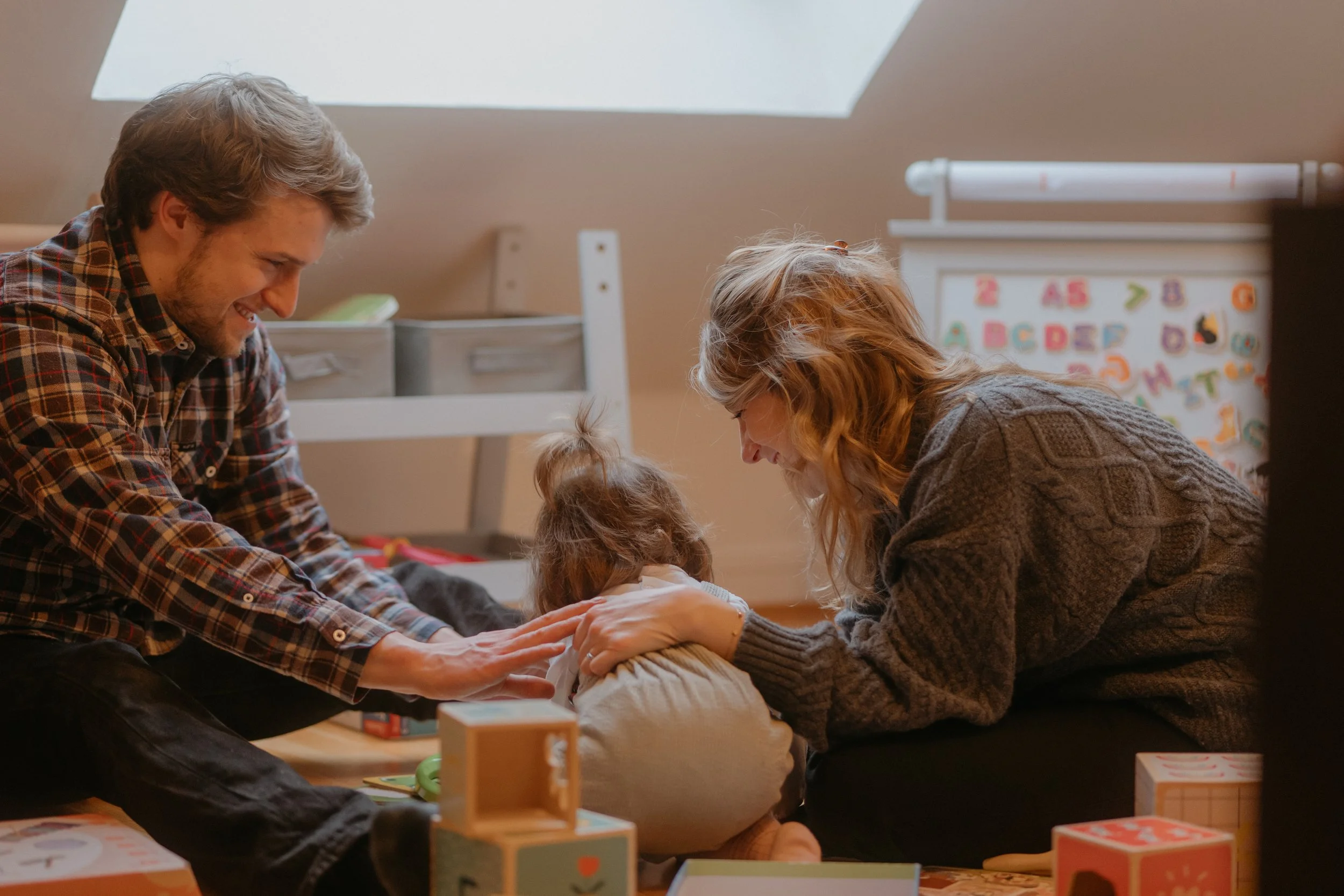 Une famille composée d'un père, d'une mère et d'une jeune fille jouent ensemble sur le sol d'une pièce lumineuse. La mère et le père sourient pendant qu'ils interagissent avec la petite.