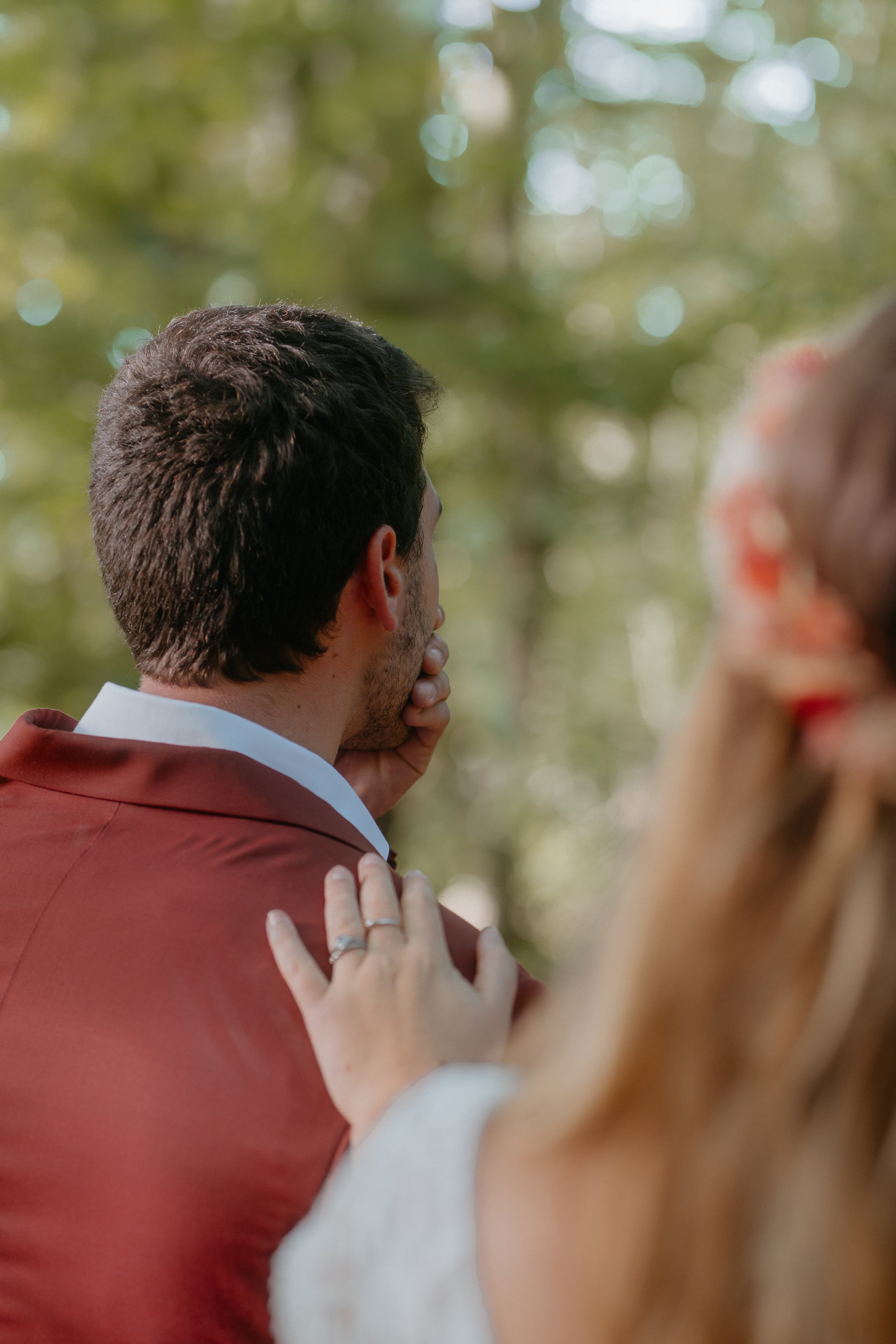 Révélation d'un couple de mariés, on voit la main de la femme avec l'alliance se poser sur l'épaule du marié en costume de mariage terracota. Le lieux est une forêt dans les Yvelines.
