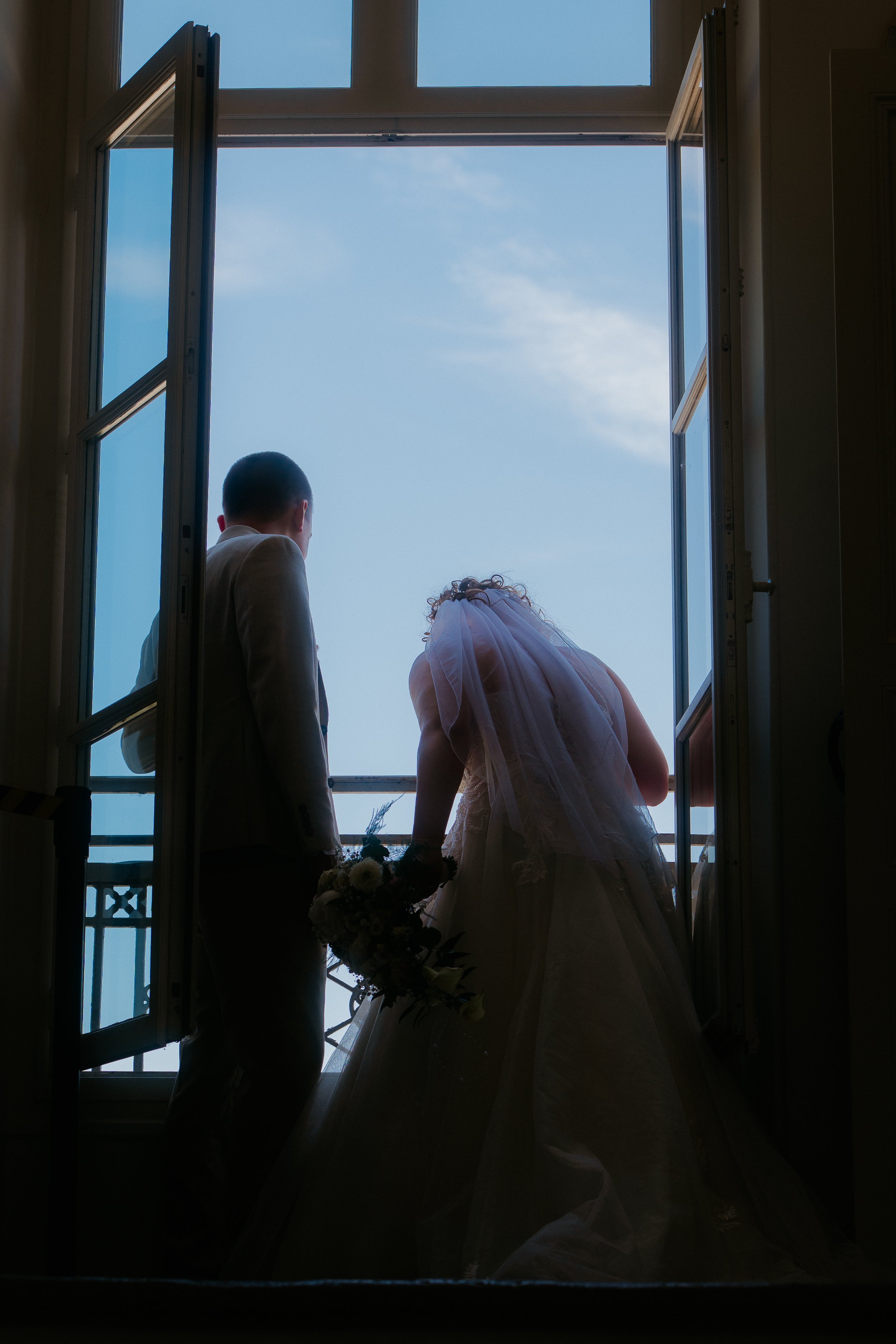Un couple de mariés, vu de derrière, debout dans une pièce ouverte en balcon, regardant vers le ciel bleu, avec la mariée tenant un bouquet de fleurs.