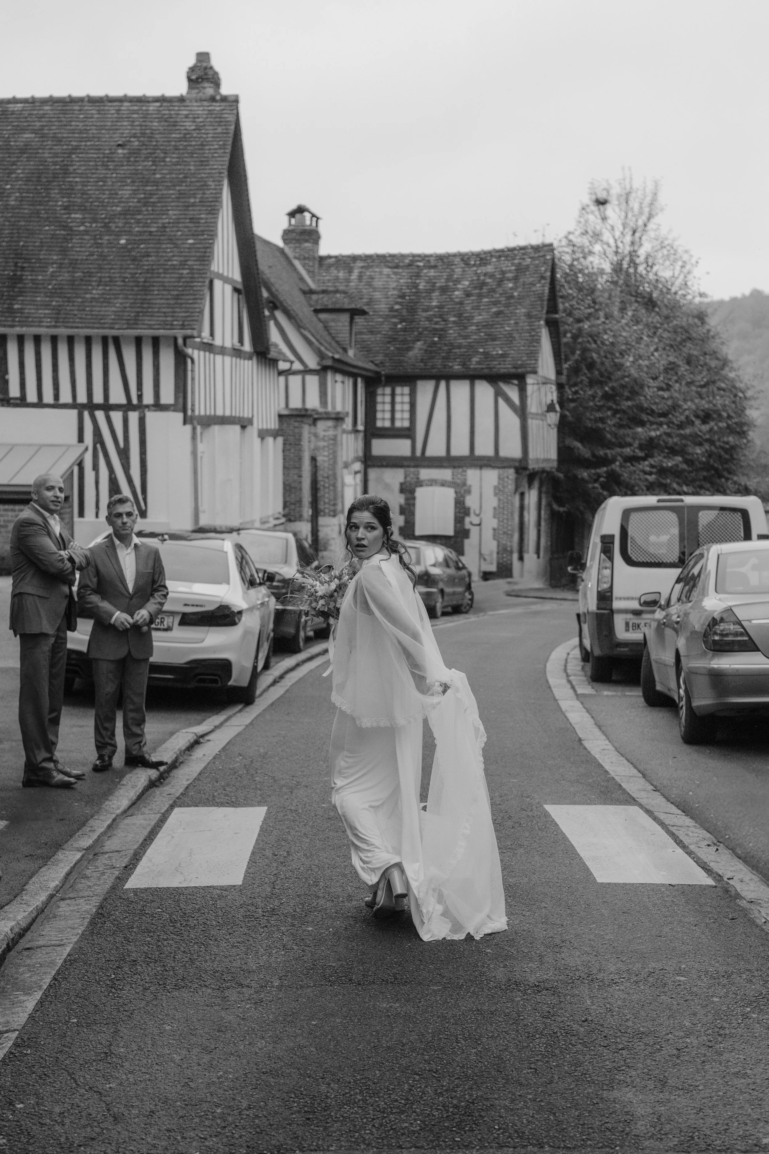 Une femme en robe de mariée traverse une rue pavée de Normandie, avec des hommes en costume qui l'observent, dans un village avec des maisons en colombages et des voitures garées le long de la rue.
