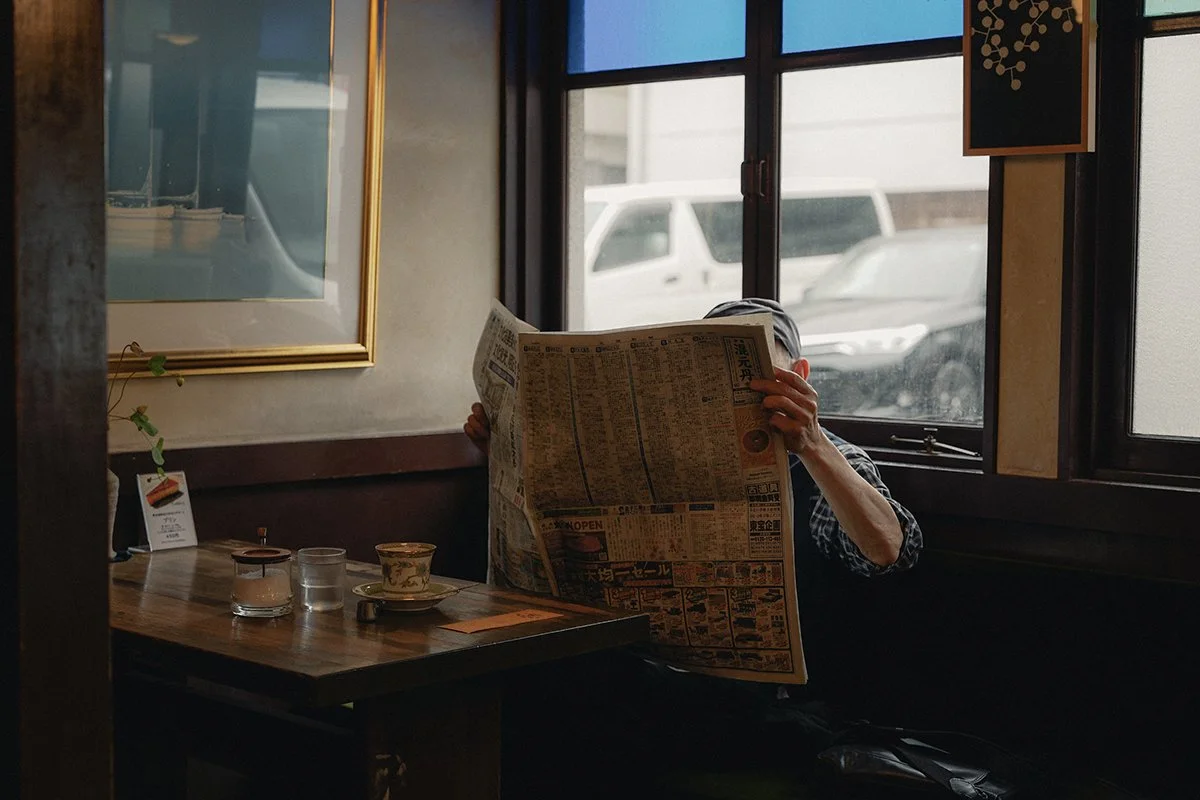 Une personne assise dans un café, lisant un journal japonais, près d'une fenêtre surgée, avec une tasse, un verre d'eau et un pot sur la table.
