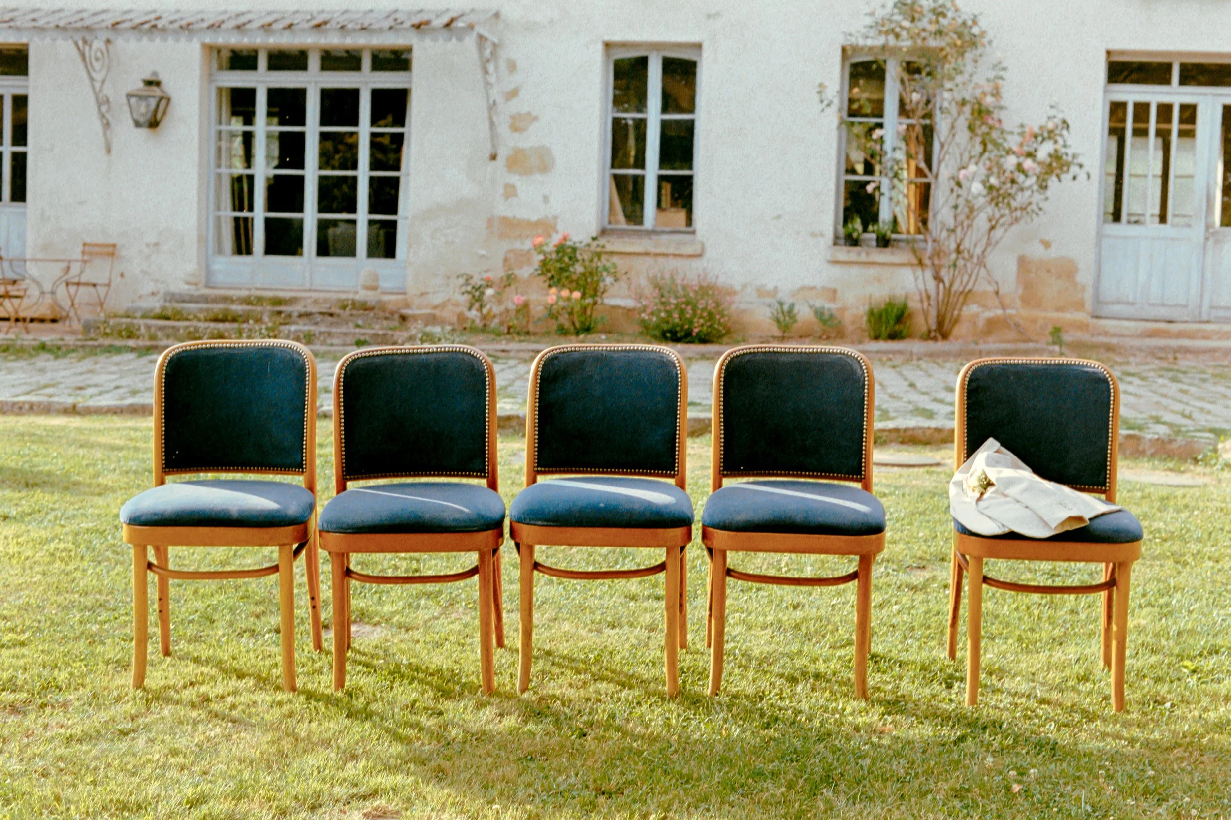 Cinq chaises en bois avec assises en tissu bleu foncé, alignées sur une pelouse verte devant une maison ancienne des Yvelines avec fenêtres et porte en bois, une veste de mariage sur la dernière chaise à droite.