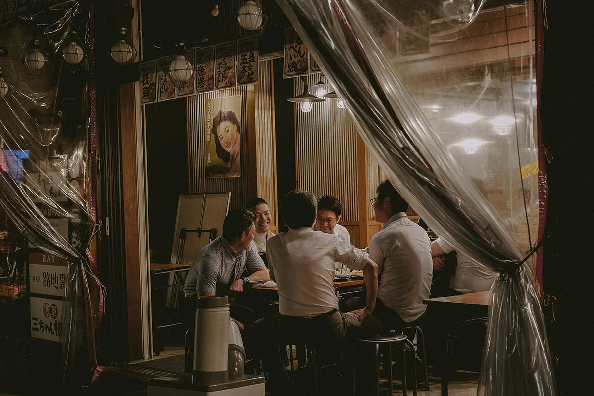 Groupe de cinq personnes assises autour d'une table dans un restaurant japonais, discutant et riant, avec des décorations traditionnelles et une peinture de femme souriante en arrière-plan.