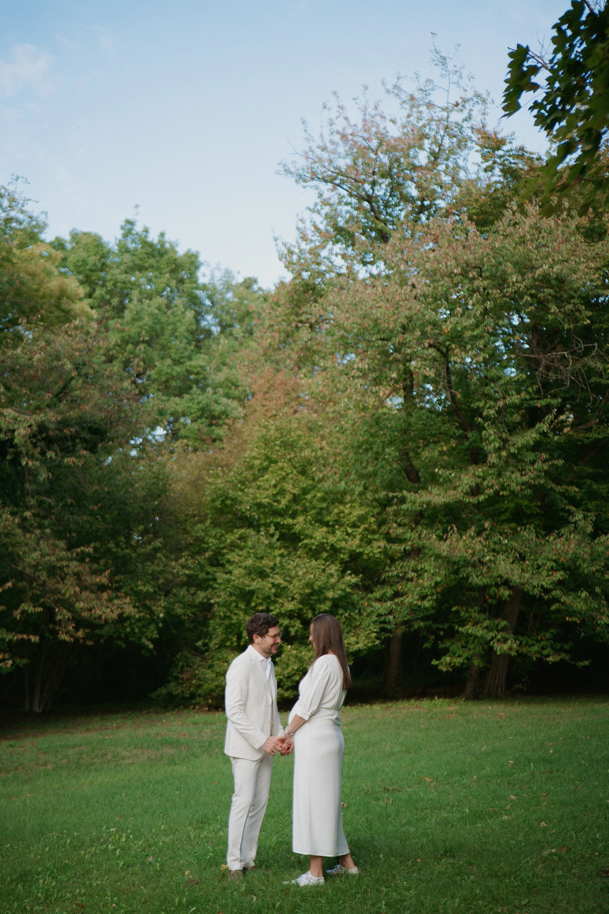 Un couple de jeune marié, en robe blanche et costume blanc, se tient la main dans un parc avec des arbres verts en arrière-plan, sous un ciel bleu clair.