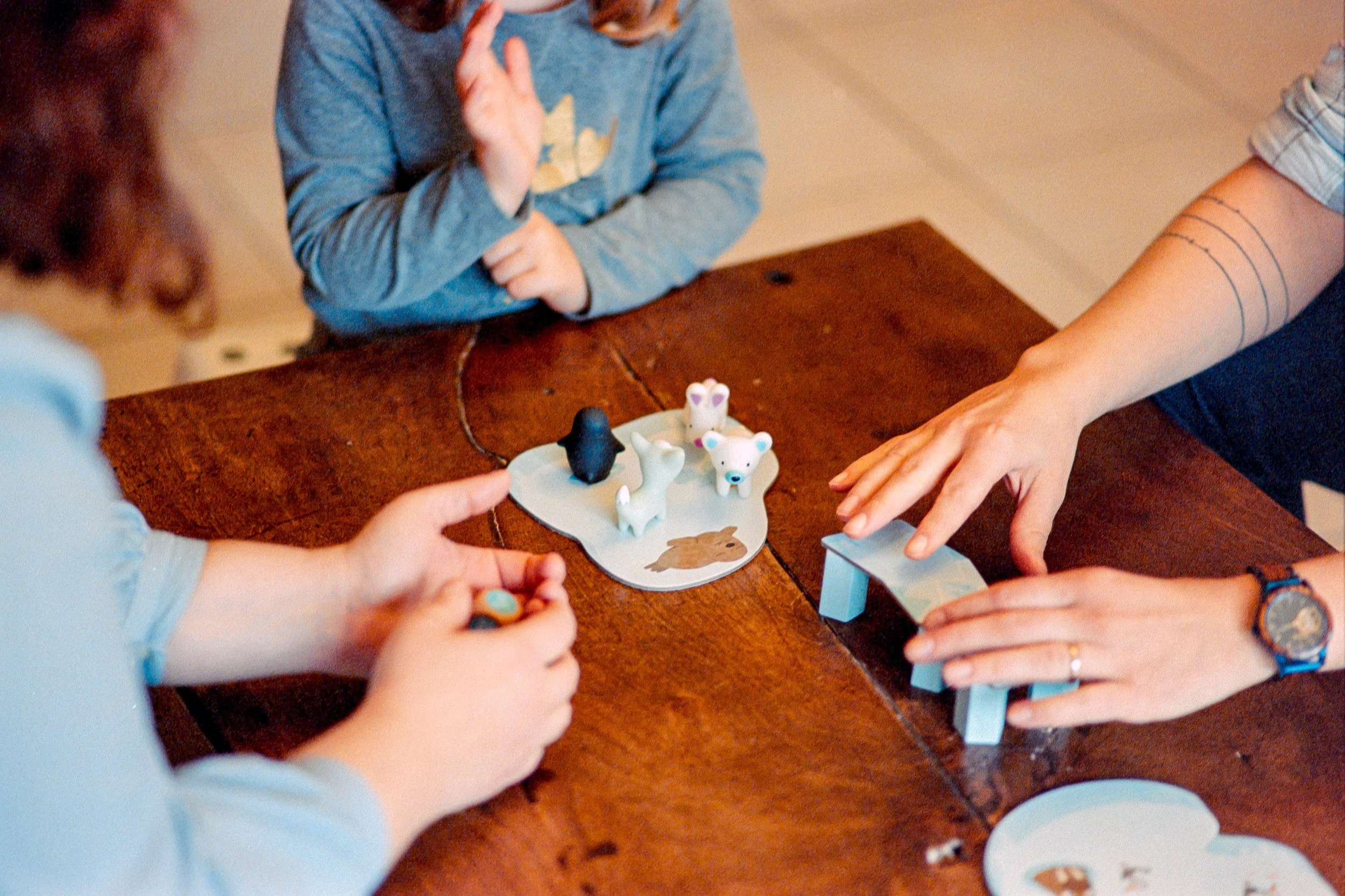 Trois personnes, d'une même famille Belle-Îloise, jouent avec des figurines d'animaux en argile ou en plastique sur une table en bois, avec une petite pièce en forme de plateforme et une carte représentant un animal.