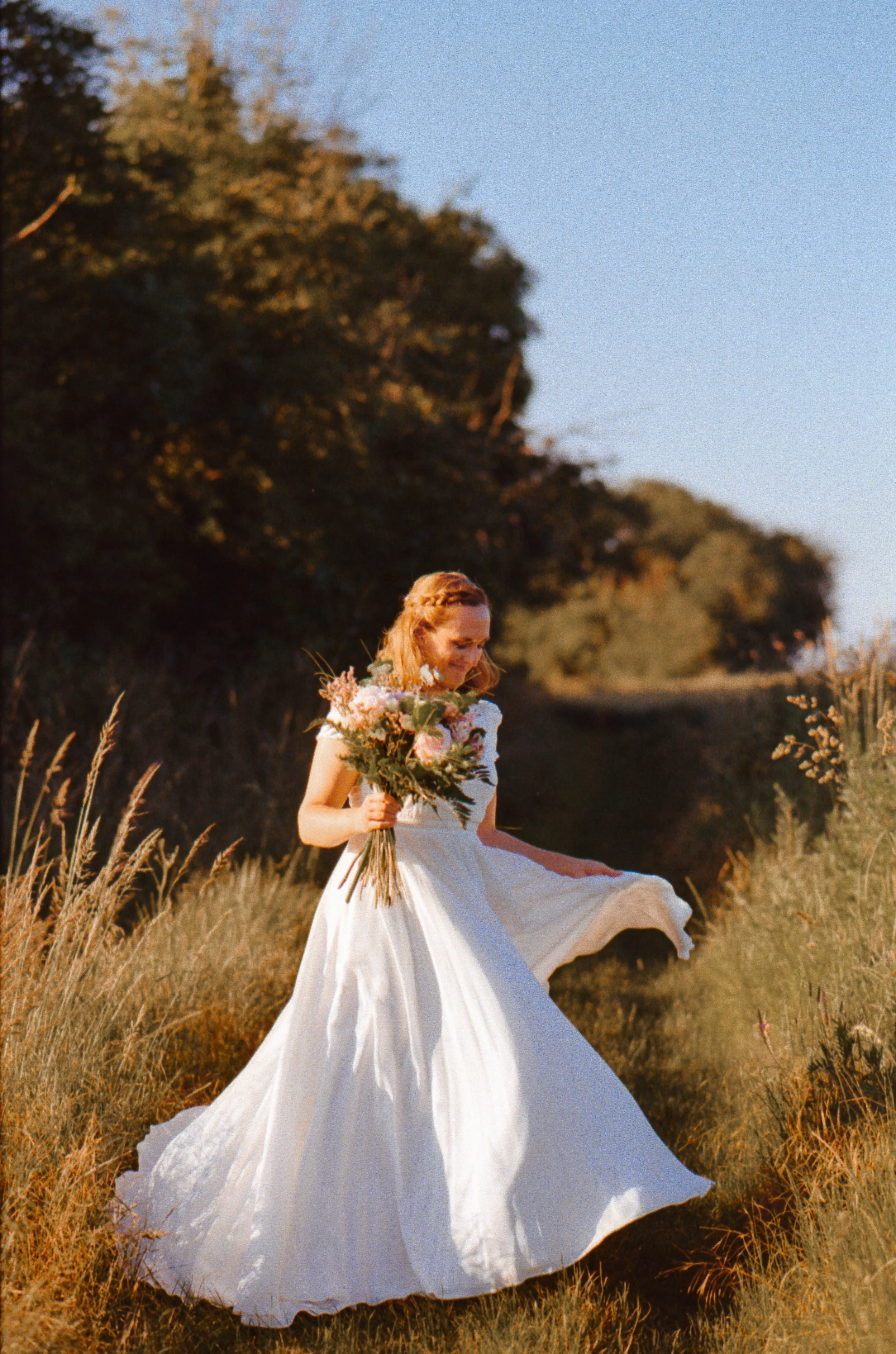 Une femme en robe de mariage tient un bouquet de fleurs dans un champ du Val d'Oise en plein air, sous un ciel clair et ensoleillé pour un beau mariage.