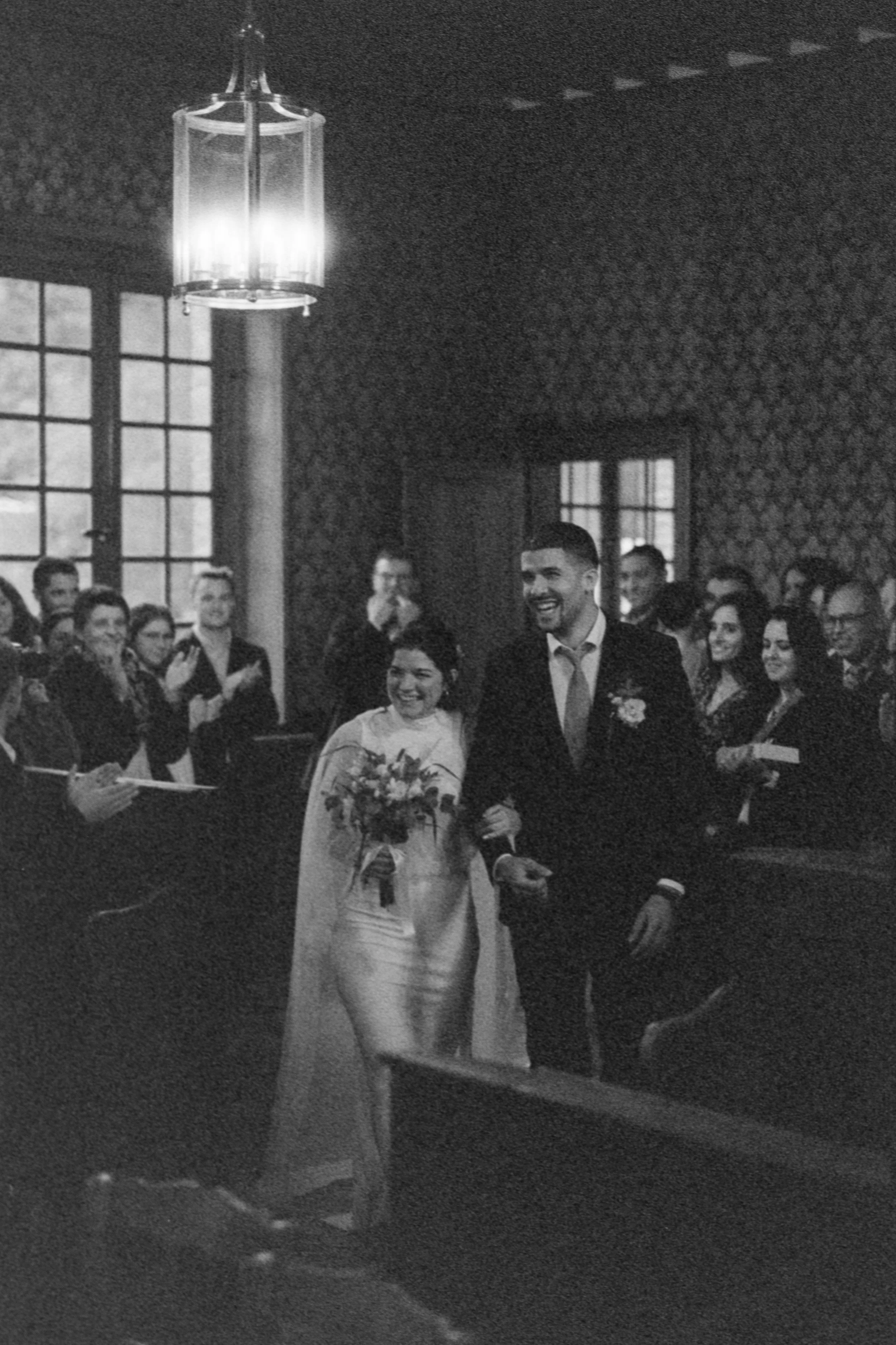 Un couple de mariés entre dans une mairie de normandie, entouré d'invités souriants lors de leur cérémonie de mariage.