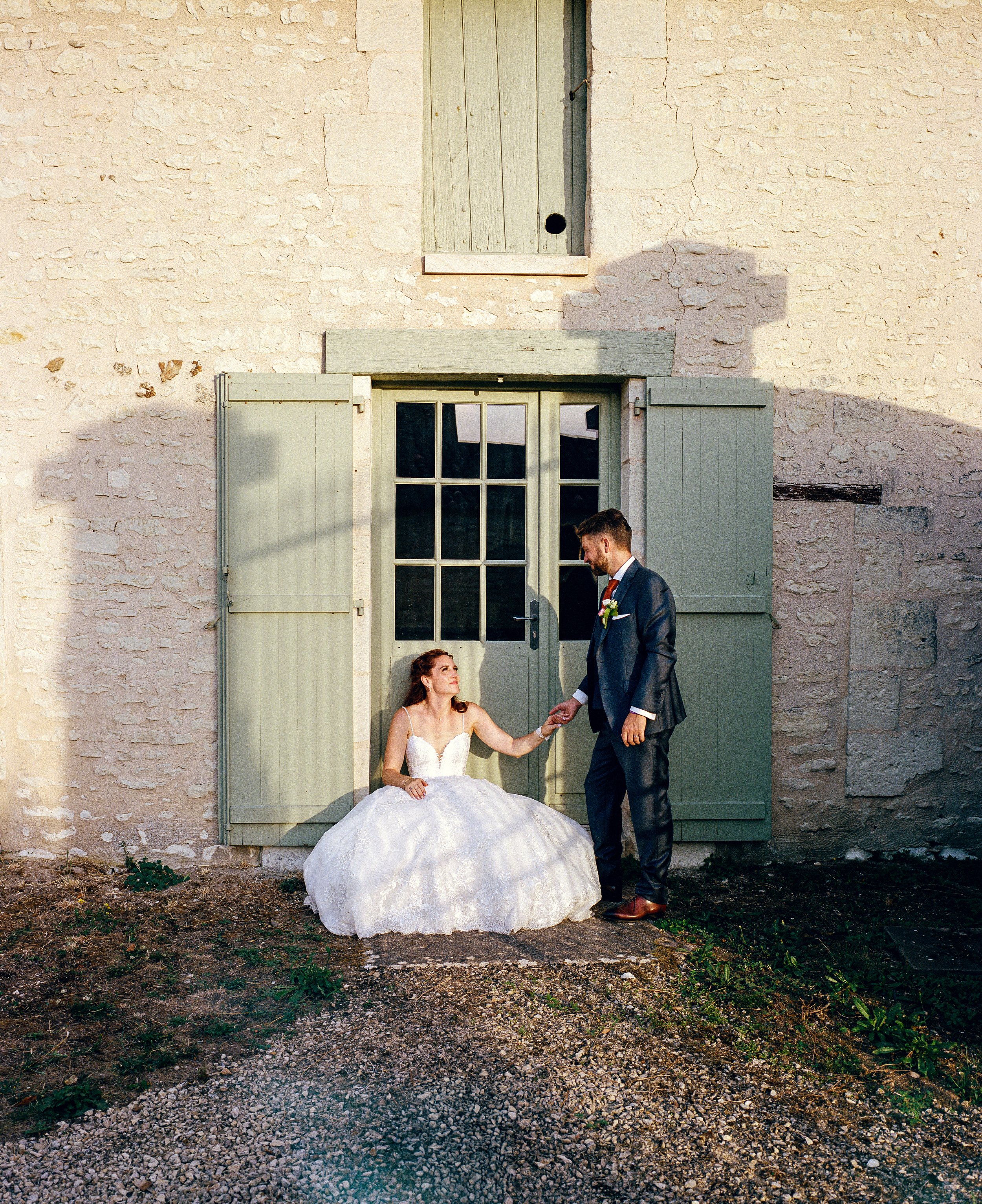 Un couple marié se tenant la main devant une maison en pierre avec des volets en bois vert pâle. La mariée est assise en robe blanche, le marié en costume sombre, dans une scène en plein air lors du coucher du soleil.