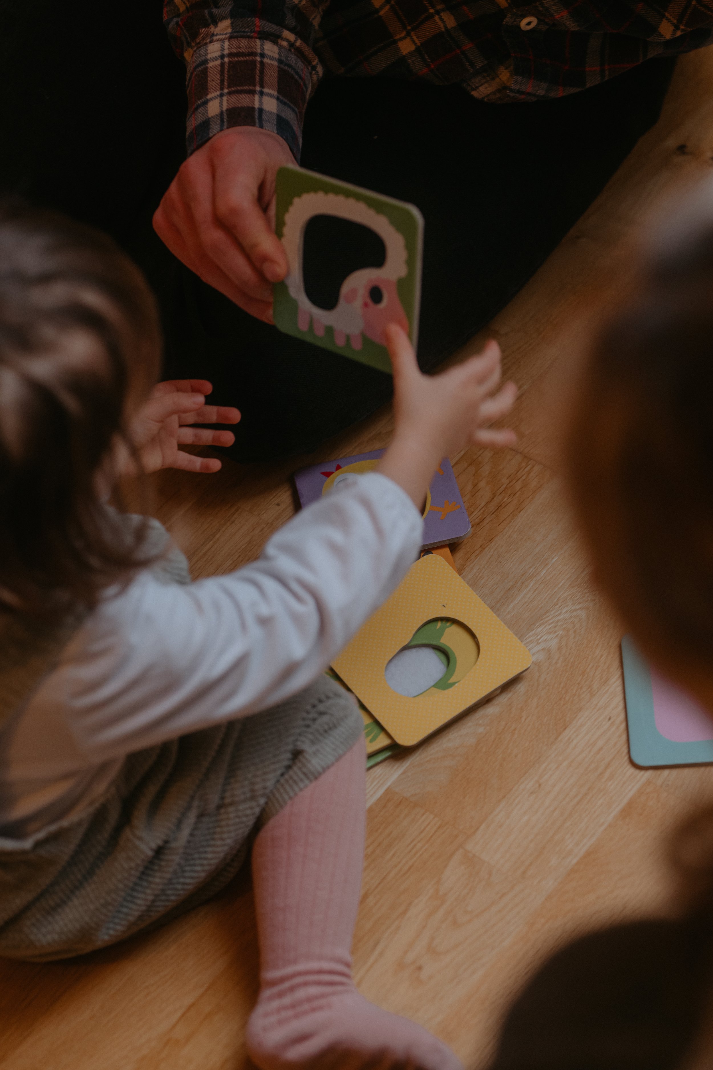 Une adulte donne un jeu de cartes illustrées d'animaux à un enfant assis sur un sol en bois, avec d'autres cartes autour.