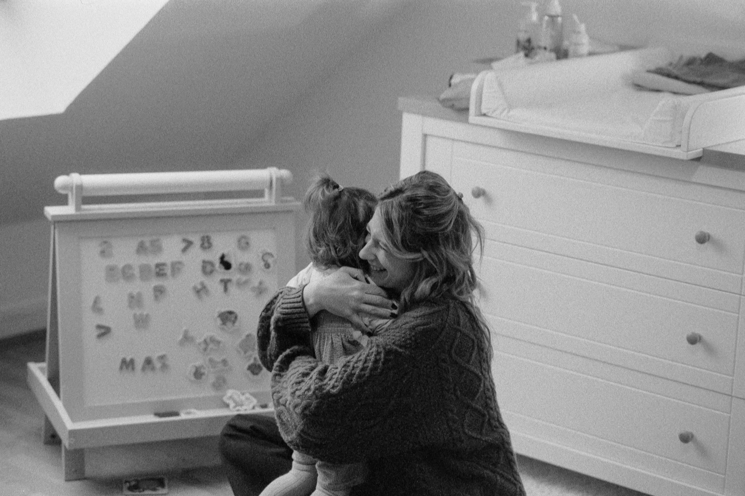 Une femme et son enfant se donnent un câlin dans une pièce, avec un tableau magnétique pour apprendre l'alphabet en arrière-plan.