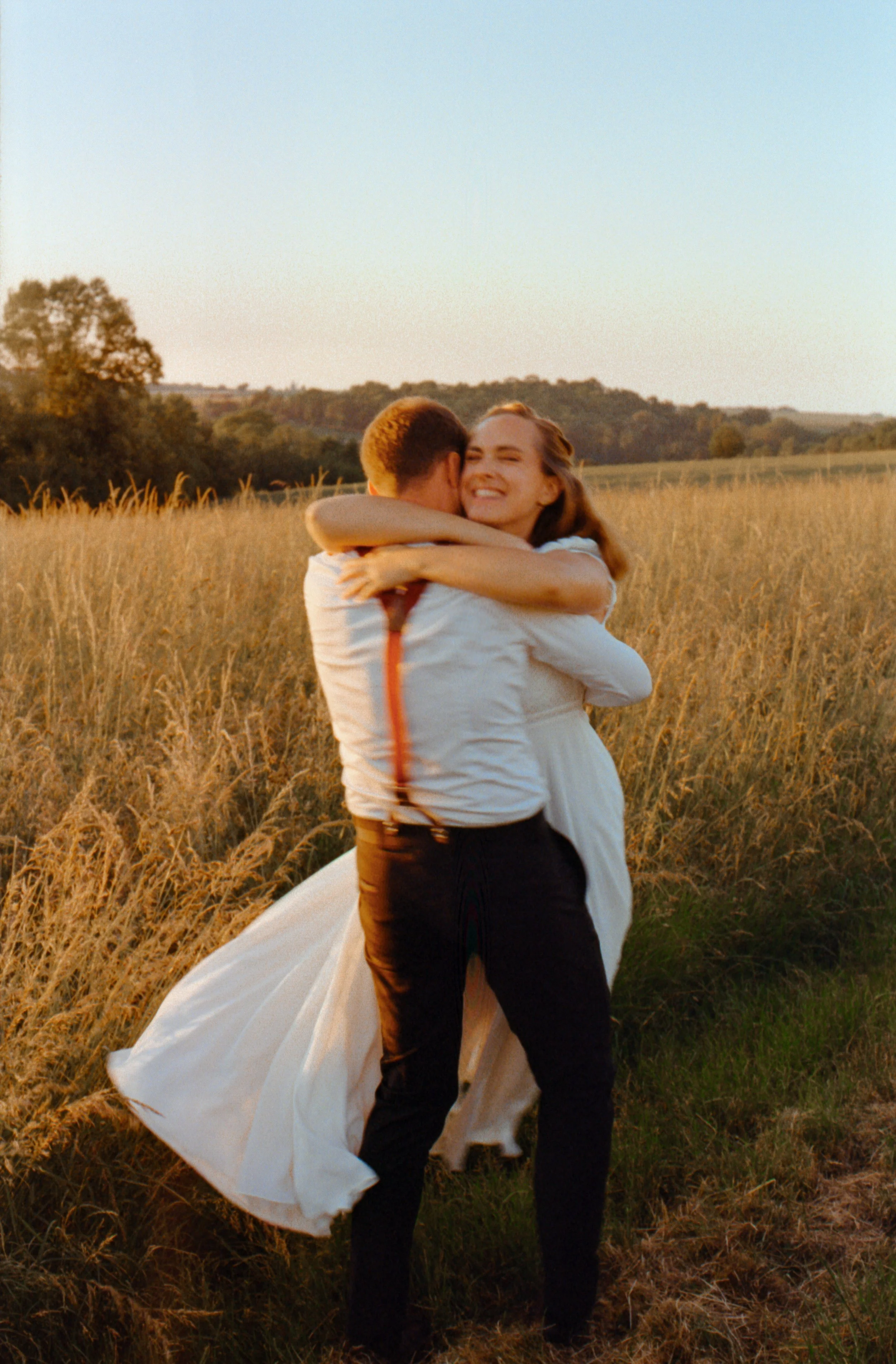Un couple de jeune marié s'embrasse dans un champ lors d'un coucher de soleil, la femme souriante portant une robe de marié et l'homme portant un costume de mariage. Ils font un tour sur eux mêmes avec une ambiance de fin de journée nostalgique.