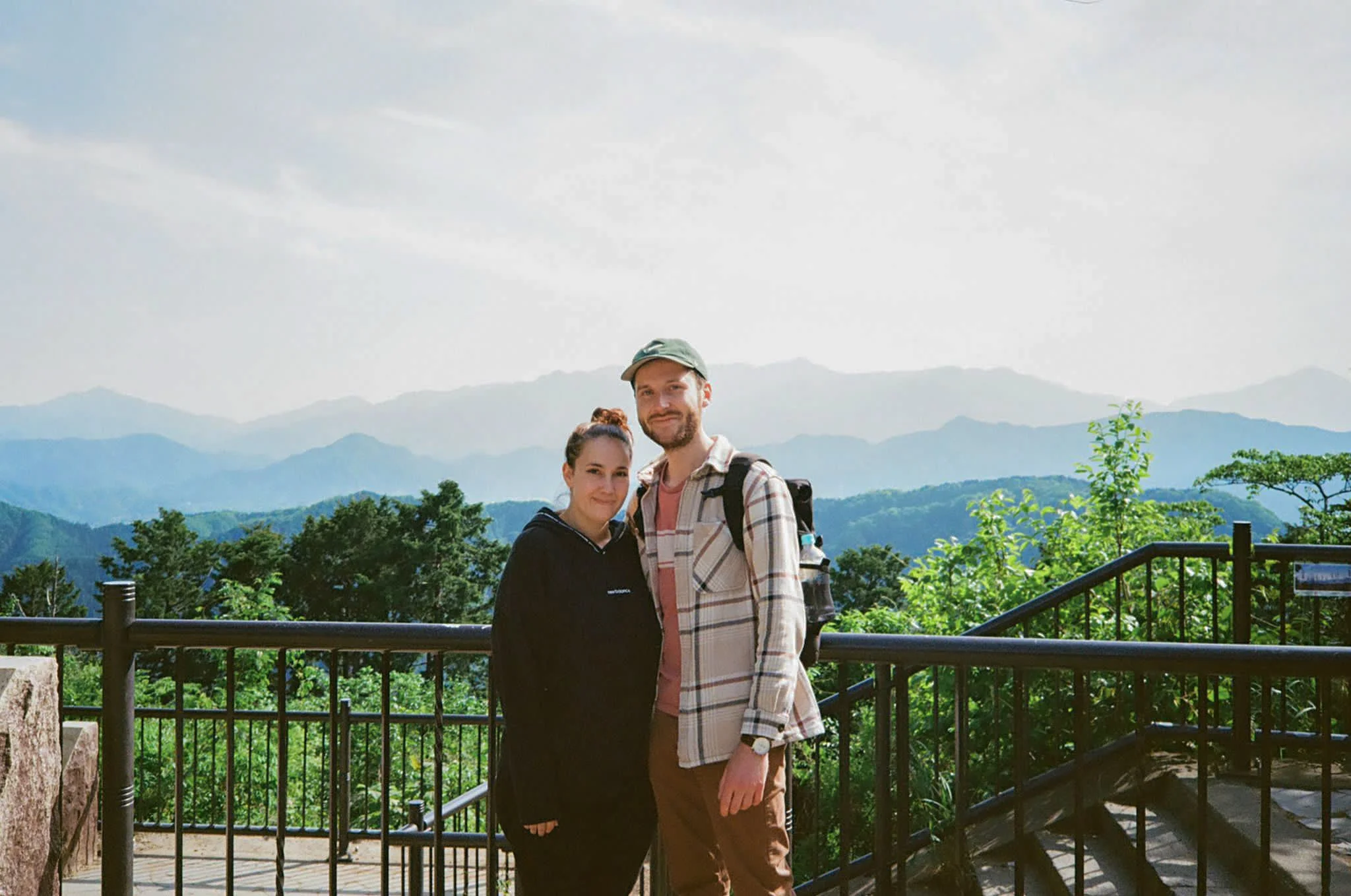 Un couple pose devant un paysage de montagnes verdoyantes avec un ciel partiellement nuageux.