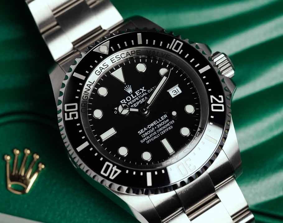 A close-up of a Rolex Sea-Dweller diving watch with a black dial and bezel, silver bracelet, and white markers, set against a green background.