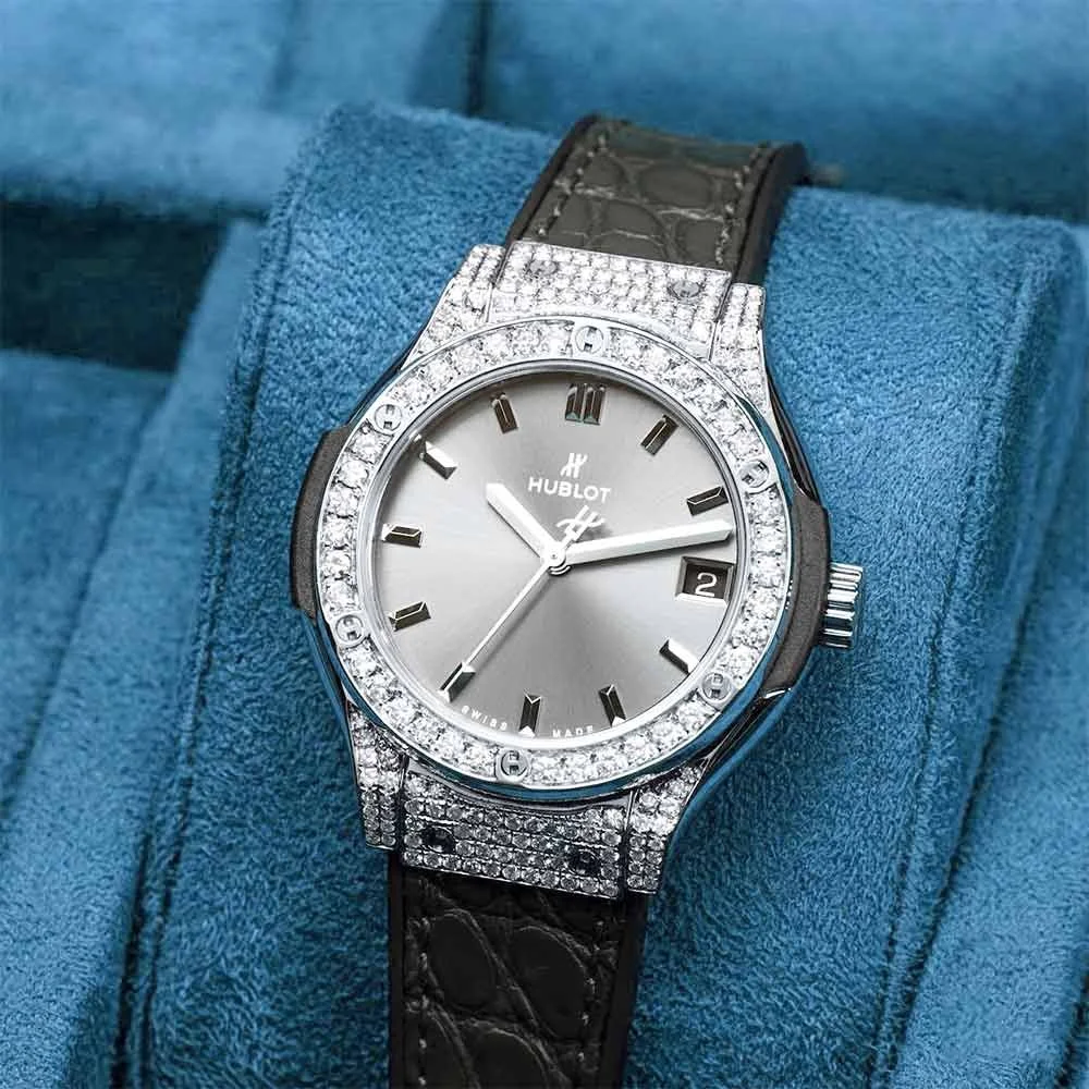 A luxury Hublot wristwatch with a silver face, diamond-studded bezel, and black strap resting on a blue fabric surface.