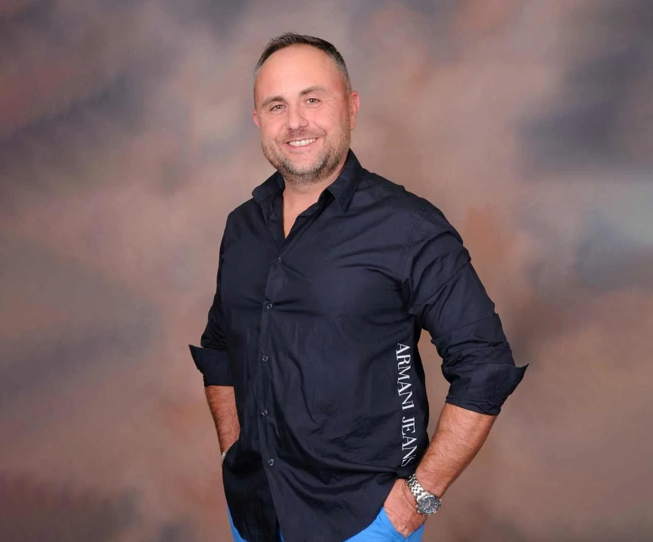 A smiling man with short hair and a beard, wearing a dark blue Armani Jeans shirt and a silver wristwatch, standing against a blurred brown and gray background.