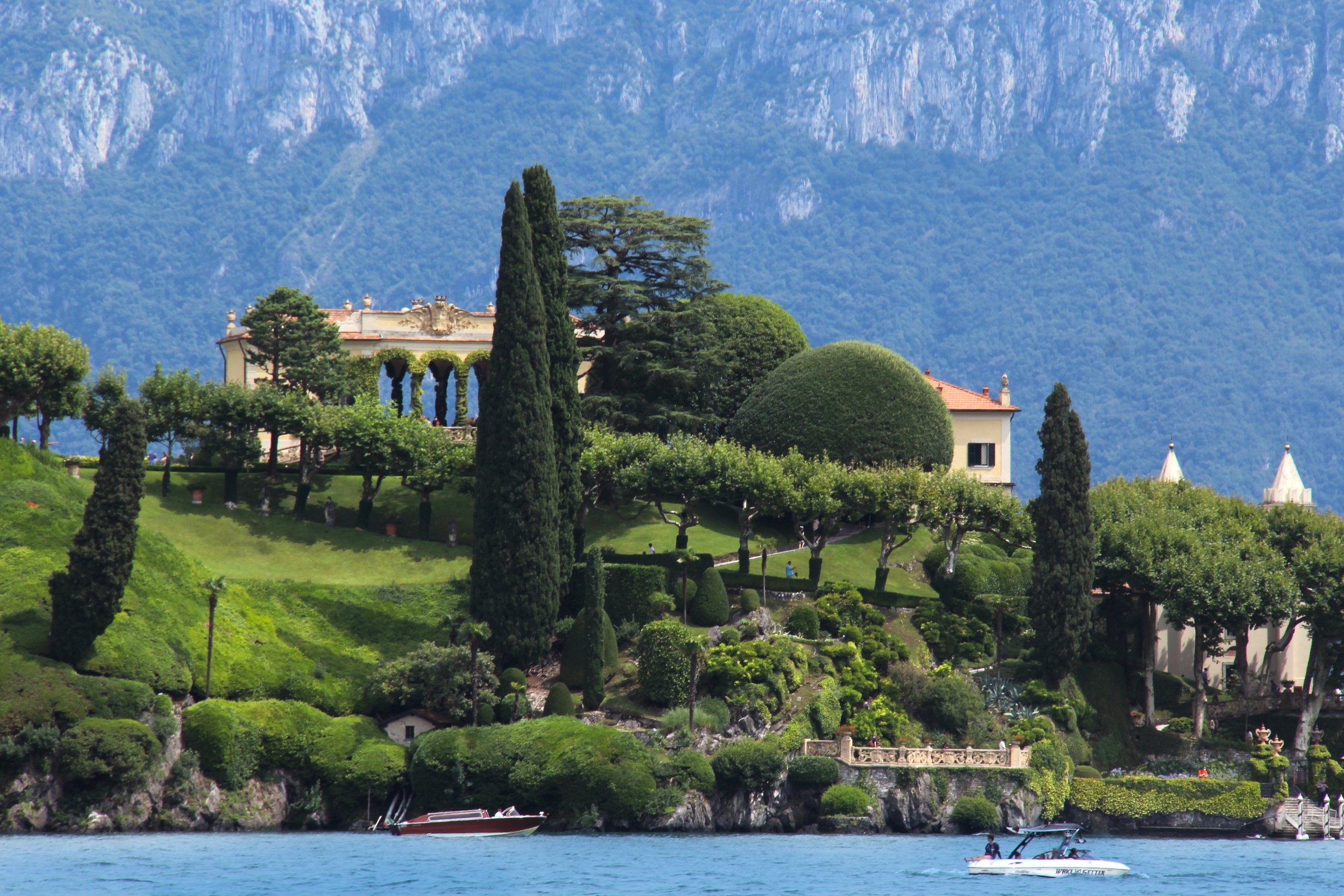 Lush hillside garden with tall cypress and trimmed trees, house with red-tiled roof, and boats on water in foreground with mountains in background.