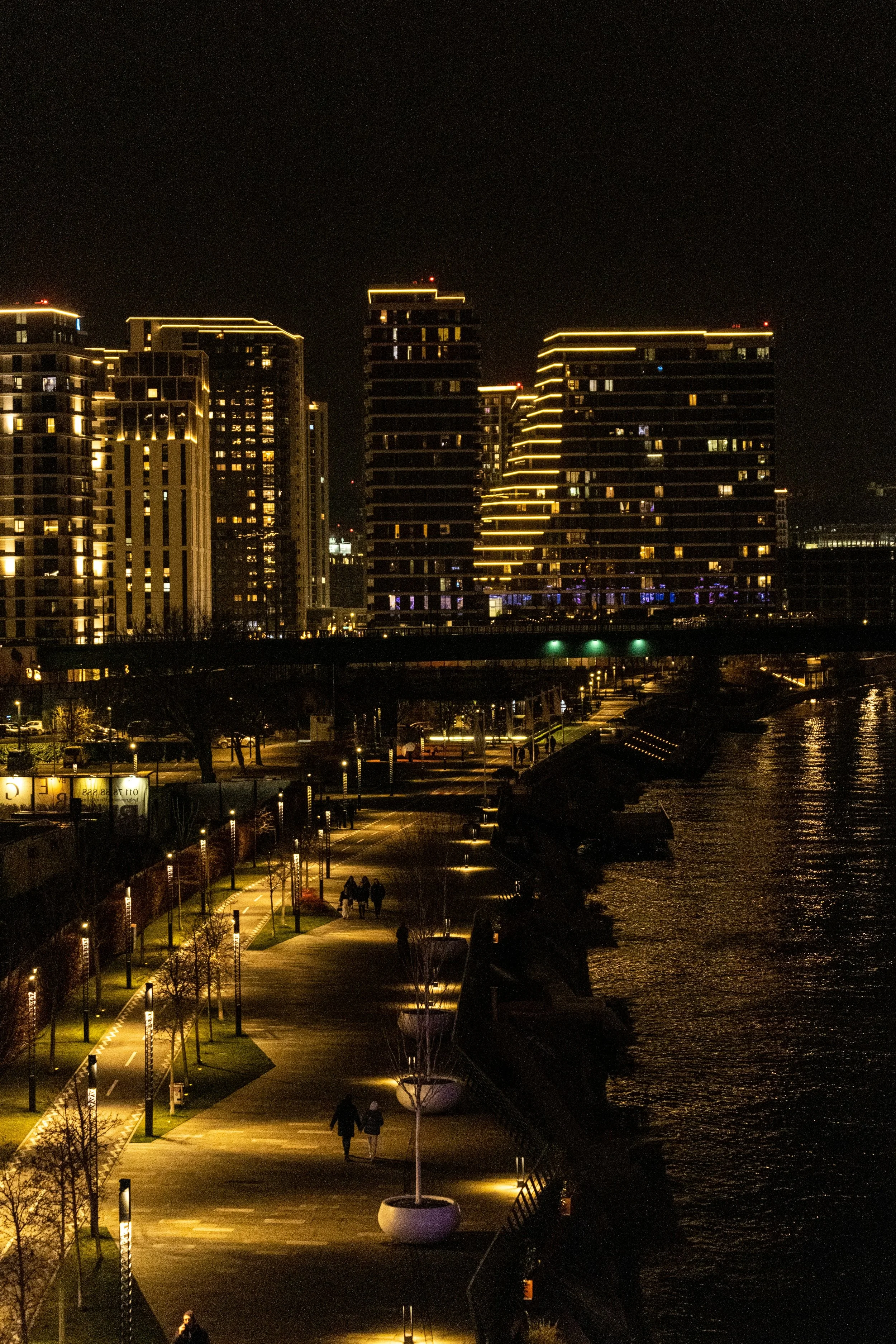 Nighttime cityscape with illuminated high-rise buildings and a waterfront promenade with people walking.