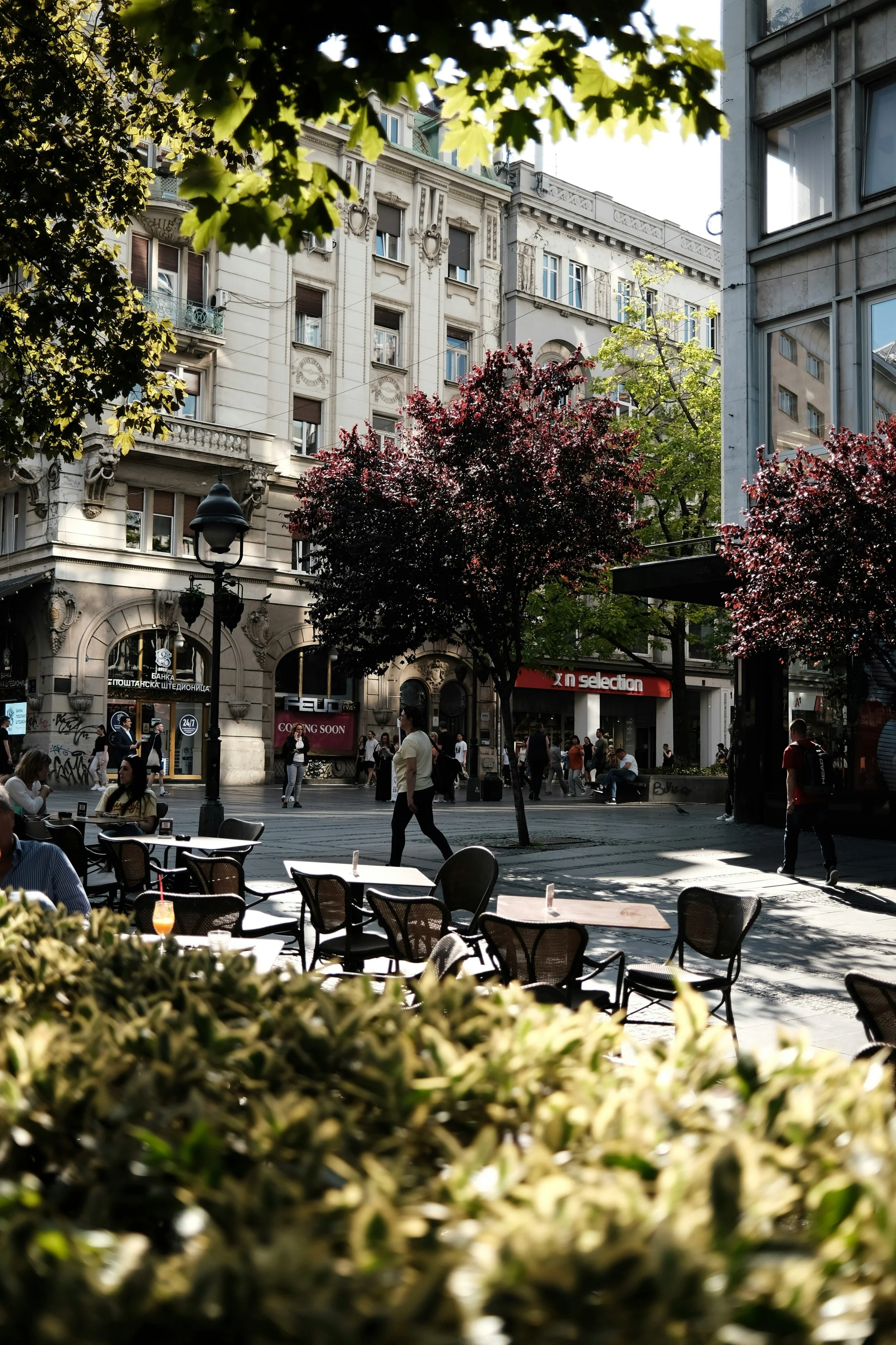 A city street scene with outdoor tables and chairs, trees, pedestrians, and historic buildings under a partly cloudy sky.