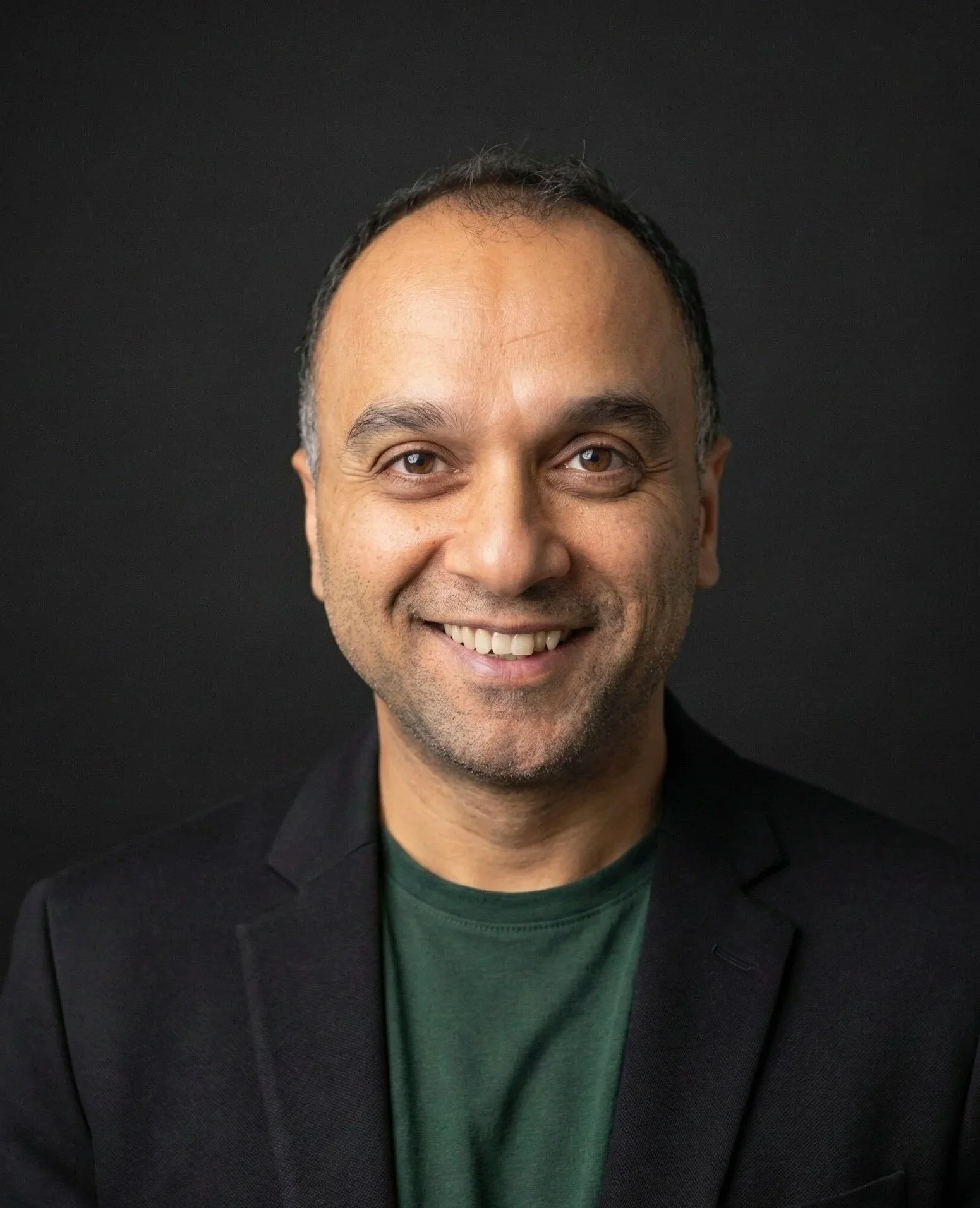 A man wearing black sunglasses, a gray suit jacket, and a black shirt, smiling at the camera against a plain beige background.