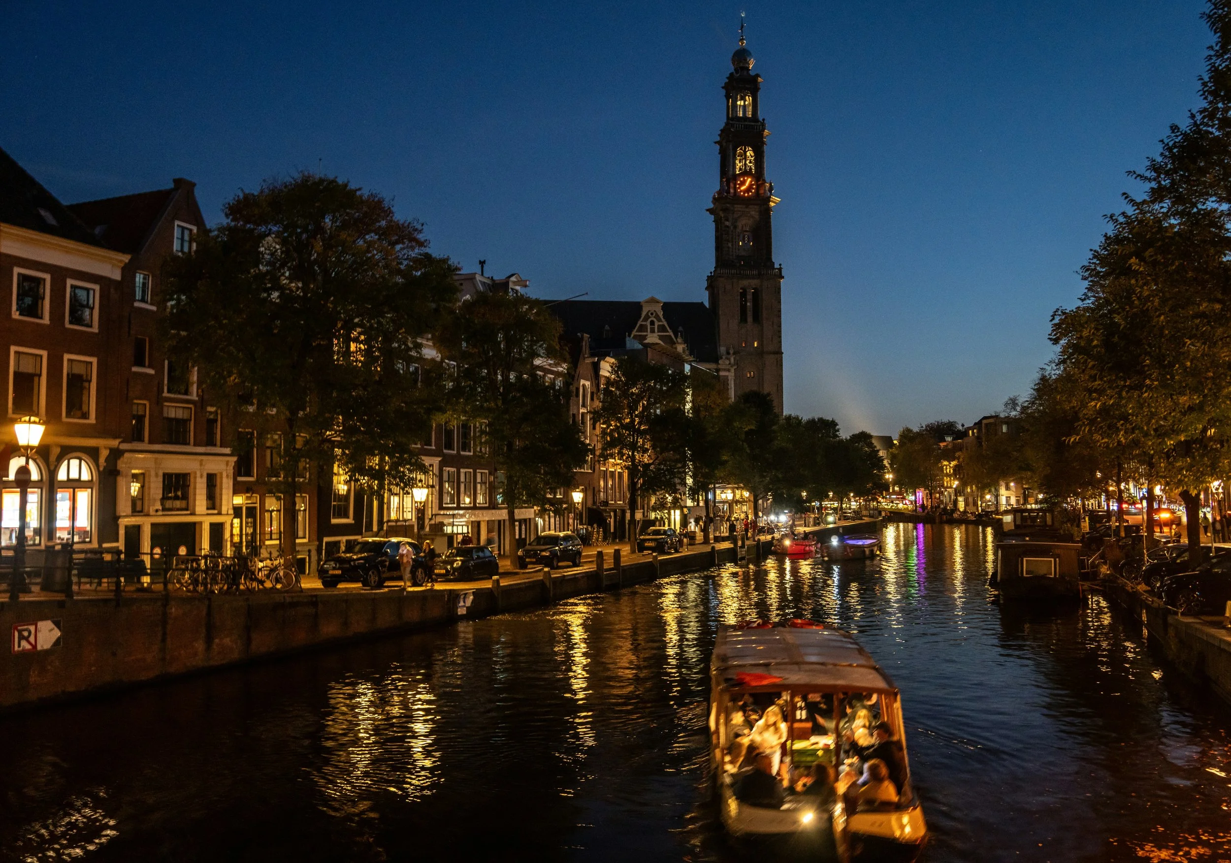 Nighttime view of a canal with a boat carrying passengers, historic buildings, trees lining the canal, and a tall clock tower in the background, illuminated by streetlights.