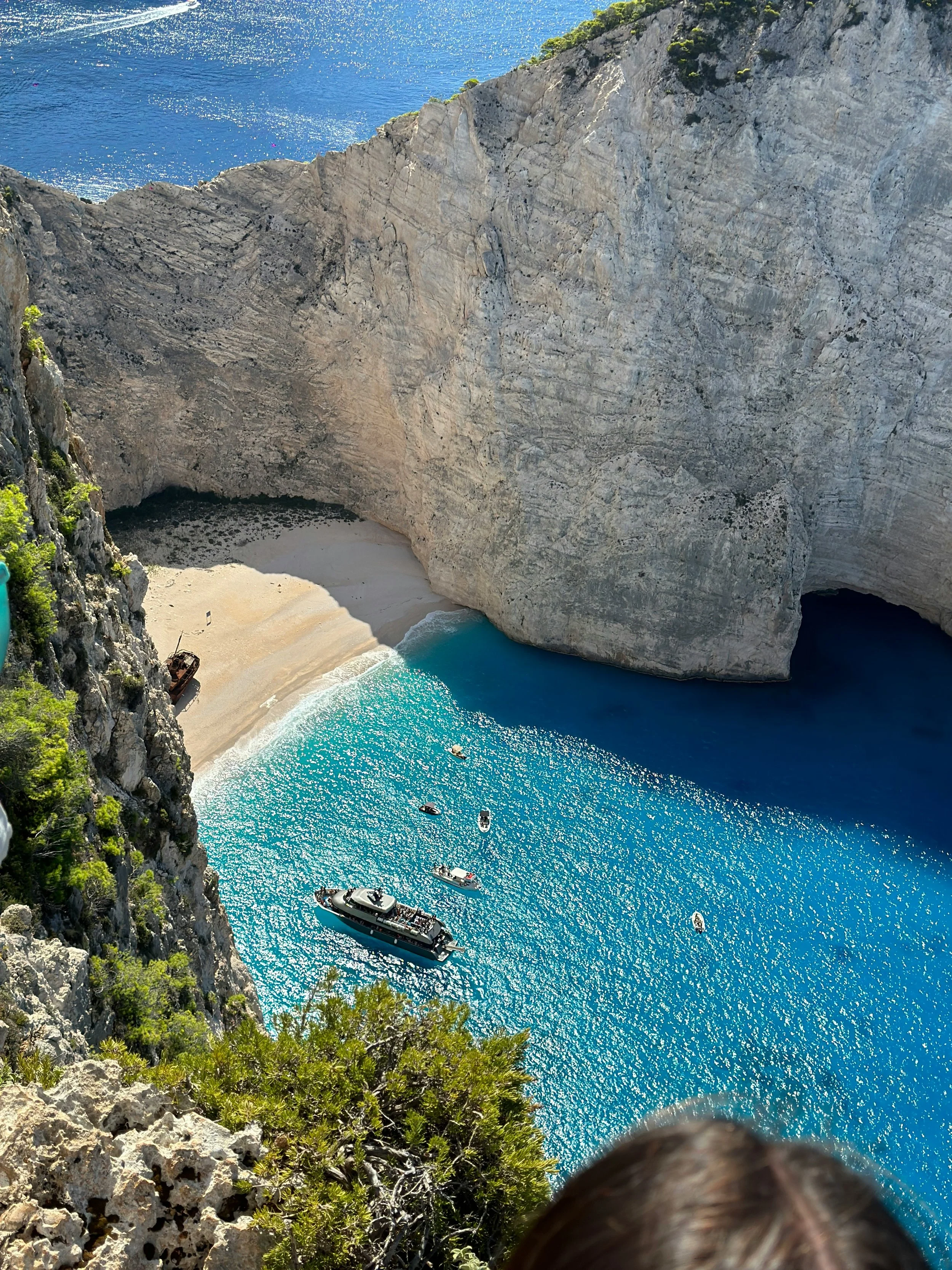High-angle view of Navagio Beach with turquoise waters, sandy cove, large limestone cliffs, and boats anchored near the shore.