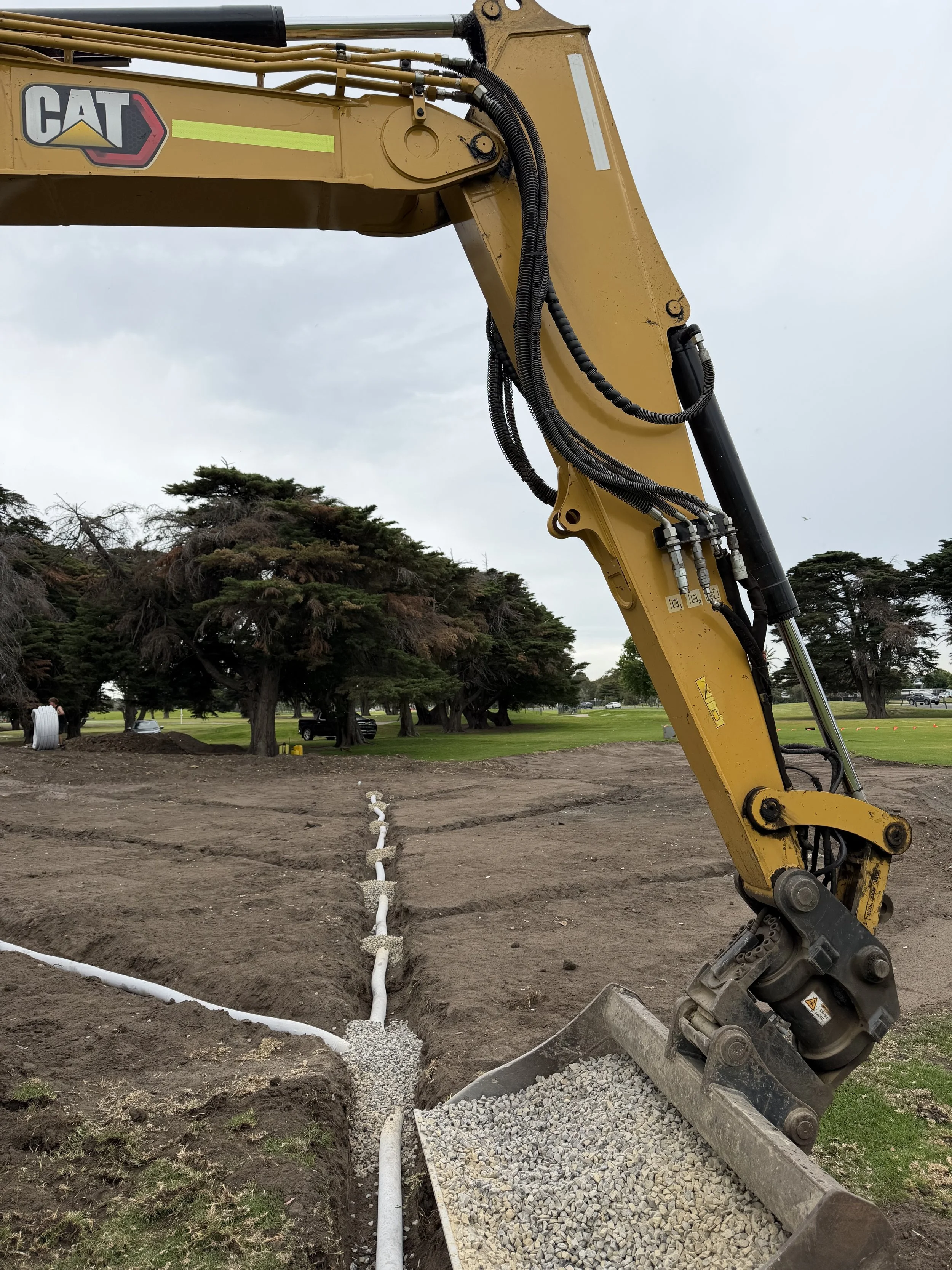Construction site with a yellow Caterpillar excavator and a trench with white plastic pipes and gravel.