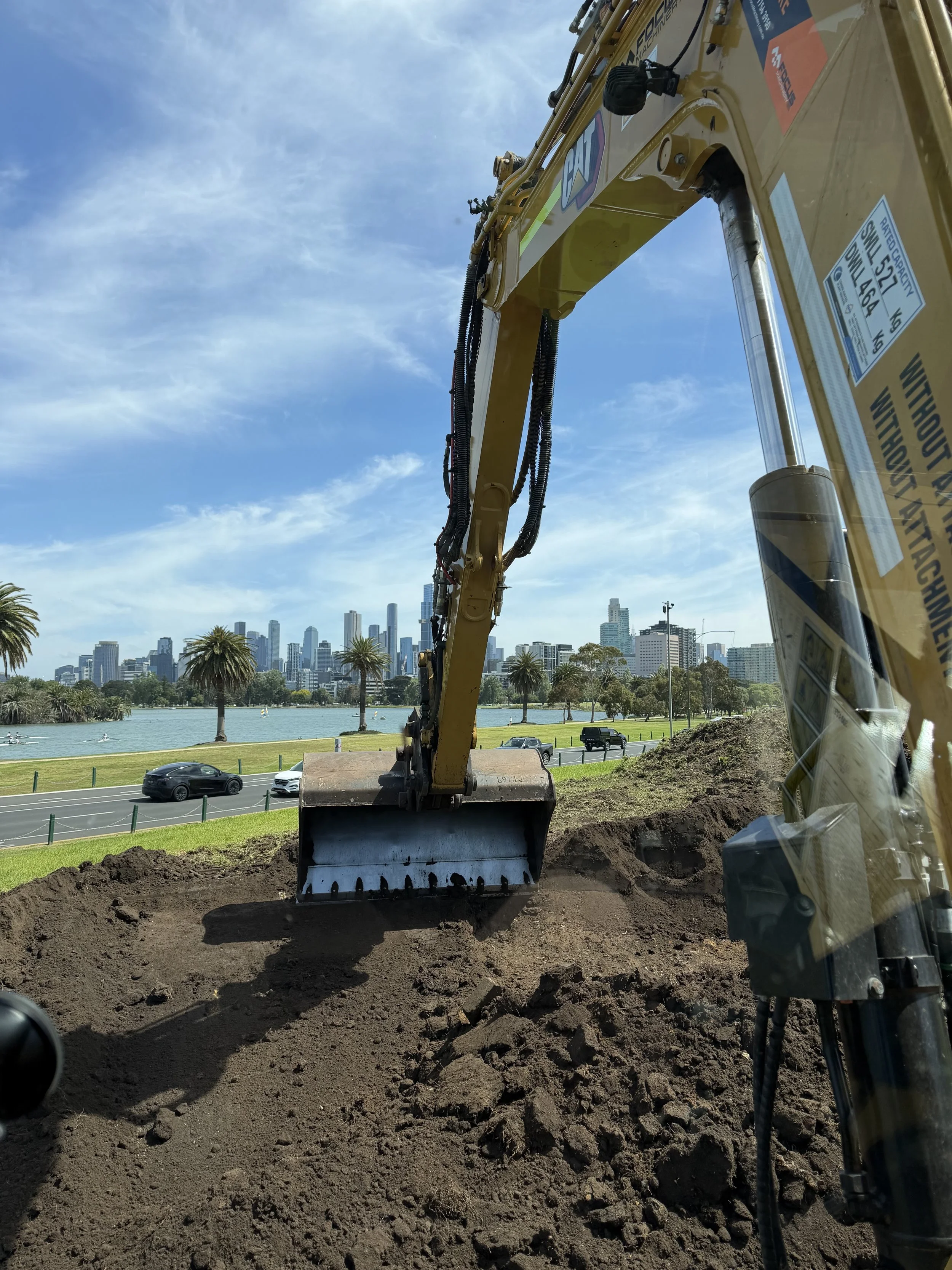 Construction excavator digging in soil near a park with palm trees and a city skyline with tall buildings in the background, under a blue sky with some clouds.