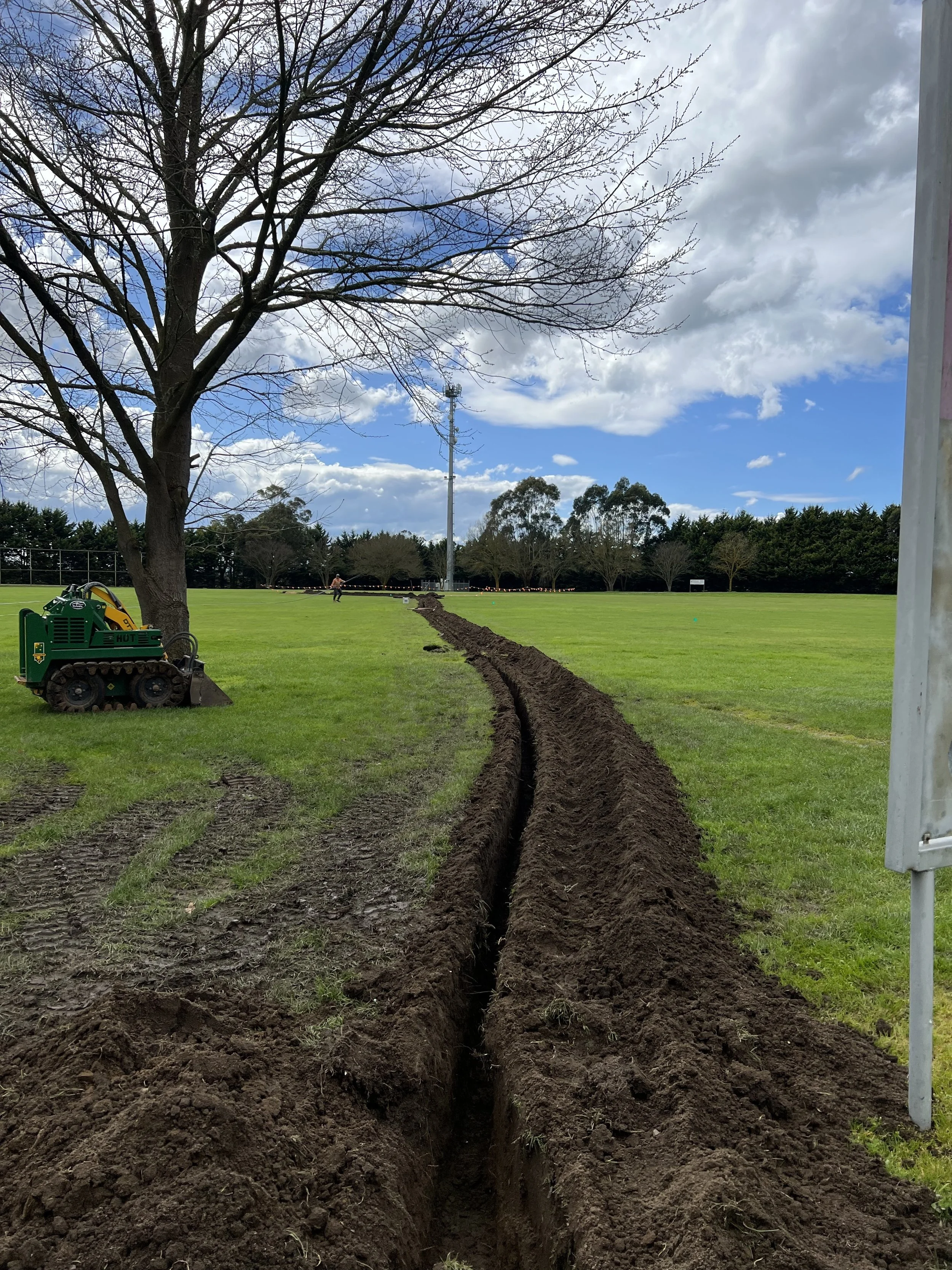 A large trench being dug in a grassy field with a small construction vehicle nearby. There is a large leafless tree and a line of trees in the background, with a partly cloudy sky above.